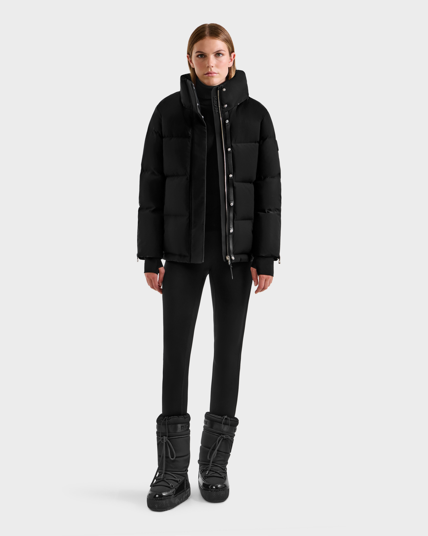A woman faces forward against a white background, wearing the Rudsak JOON H - 8125586 Black insulated quilted jacket. She pairs it with black pants and boots, her hair pulled back and expression neutral.