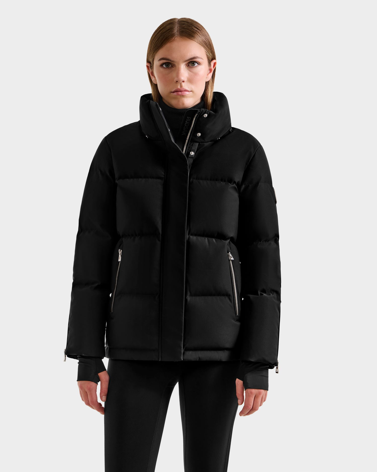 A woman faces forward wearing the Rudsak JOON H - 8125586 Black women’s down jacket with a high collar and zippered pockets, paired with black pants, against a plain light gray background.