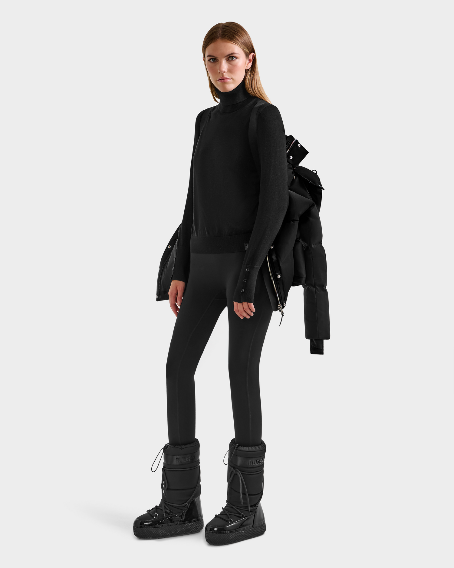A woman stands against a plain background wearing the Rudsak JOON H - 8125586 Black insulated quilted jacket draped over her shoulders, along with a black turtleneck, black leggings, and black winter boots.