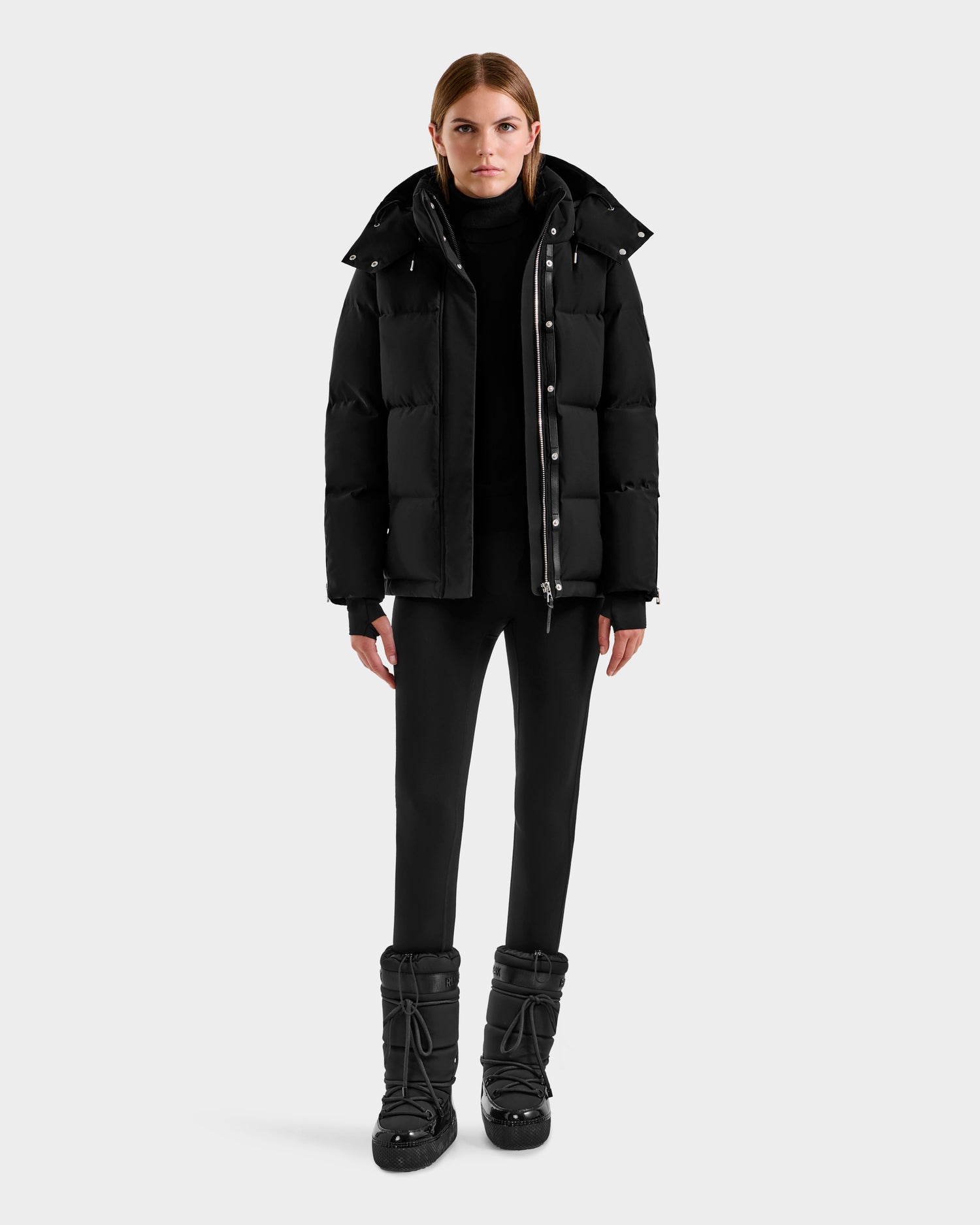 A woman faces forward against a light gray background, wearing the Rudsak JOON H - 8125586 Black insulated quilted jacket, paired with a black turtleneck, black pants, and black snow boots.