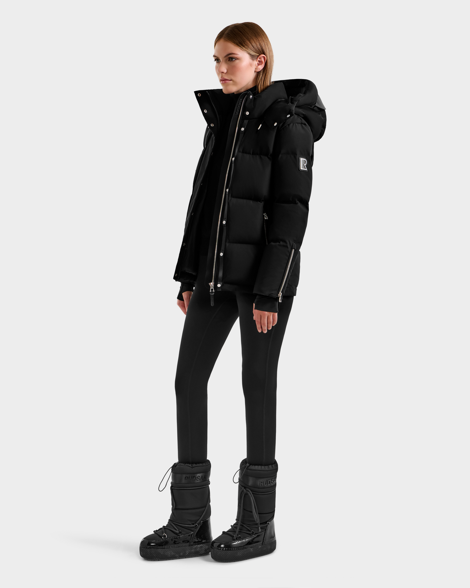 A person stands facing left wearing the JOON H - 8125586 Black insulated quilted jacket with a hood by Rudsak, paired with black pants and black lace-up winter boots, against a plain light gray background.