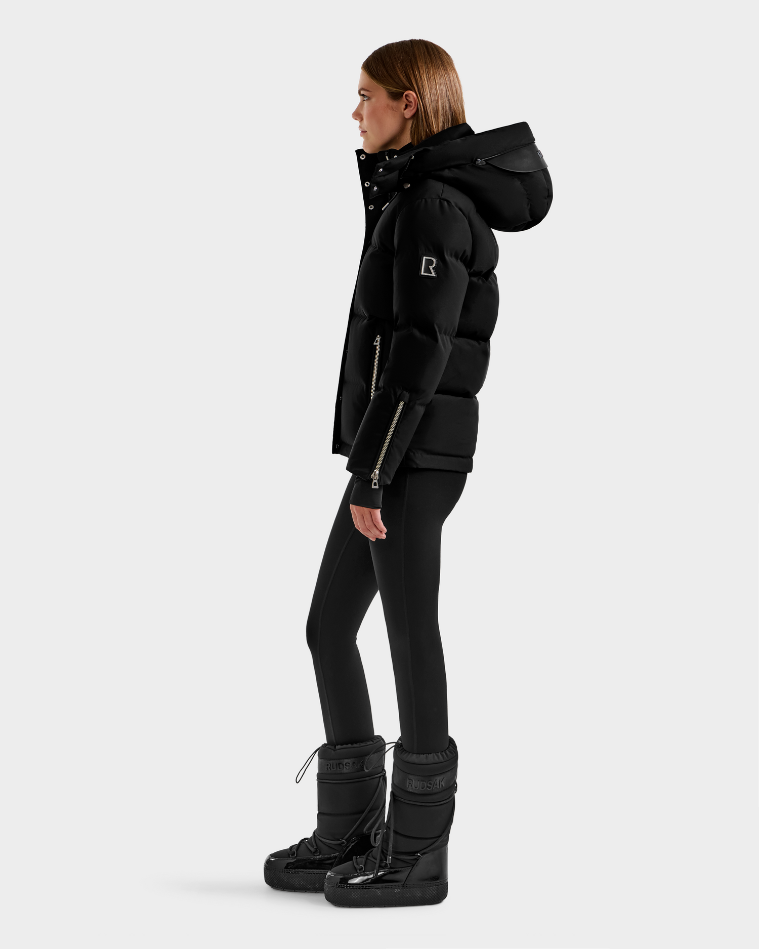 A woman in side profile stands against a light background, wearing the Rudsak JOON H - 8125586 Black down jacket with a hood, paired with black leggings and snow boots—her stylish winter coat look is perfect for cold weather.
