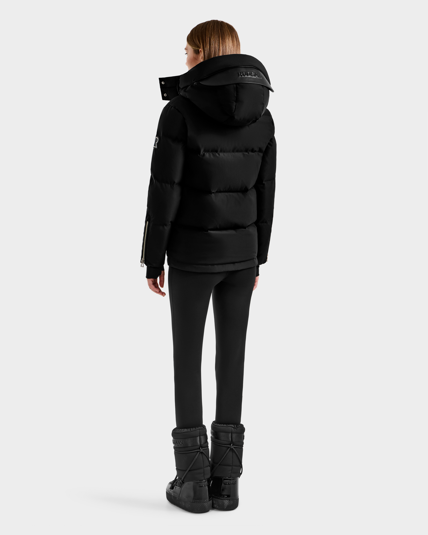 A person with straight brown hair, wearing the Rudsak JOON H - 8125586 Black insulated quilted jacket, black leggings, and black winter boots stands facing away against a plain light gray background.