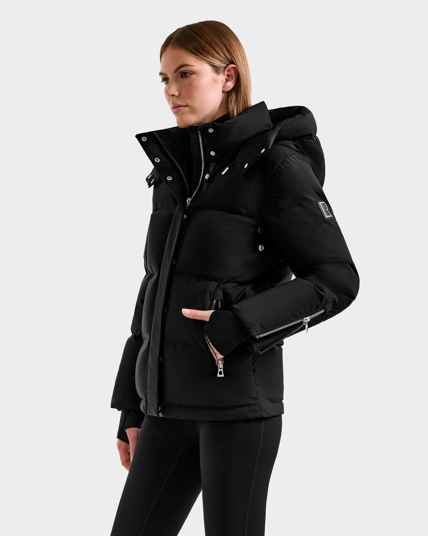 A woman with straight brown hair wears the Rudsak JOON H - 8125586 Black women’s down jacket, featuring a hood, zippered pockets, and silver hardware. She stands against a light gray background highlighting its lightweight warmth.