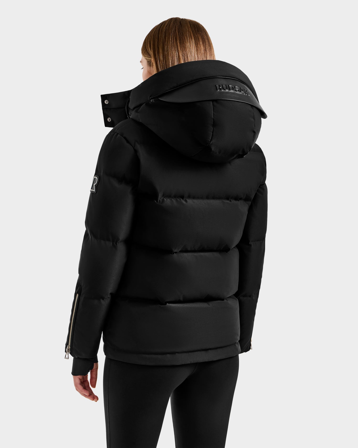 A woman with straight, light brown hair is seen from the back wearing the Rudsak JOON H - 8125586 Black insulated quilted jacket with a large hood and black pants, set against a plain light gray background.