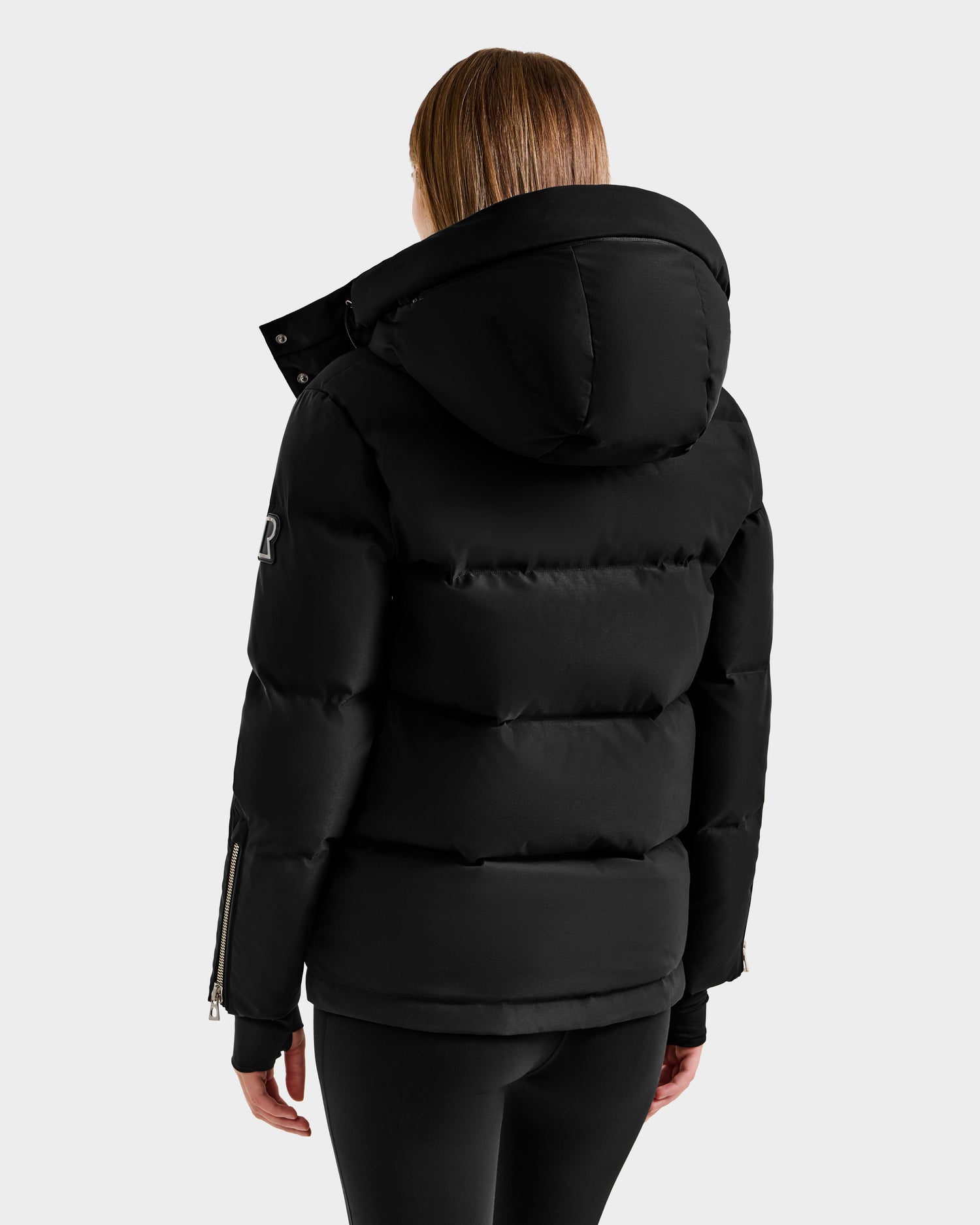 Seen from the back against a plain light background, a person with straight, light brown hair wears Rudsak’s JOON H - 8125586 Black insulated quilted jacket with a large hood and zippered sleeves, paired with black pants.