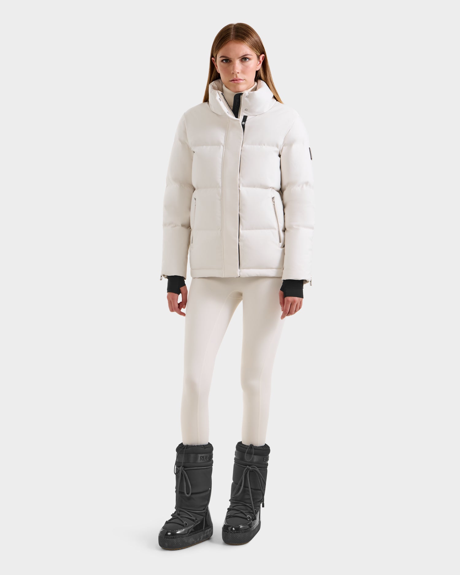 A woman faces forward against a white background, wearing the Rudsak JOON H - 8125586 Vanilla quilted winter jacket with a recycled shell, paired with white leggings and black winter boots.