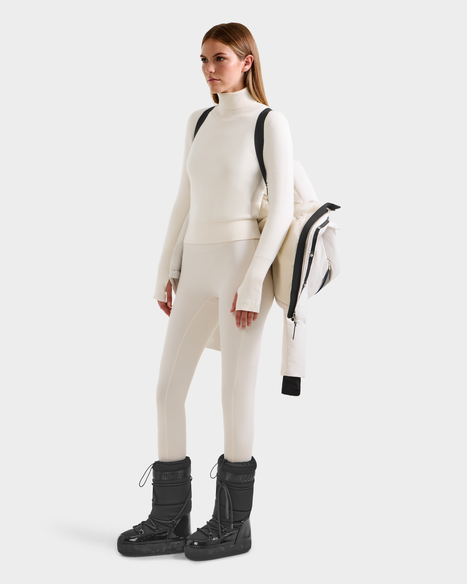 A woman with straight light brown hair faces left, wearing the Rudsak JOON H - 8125586 Vanilla women’s down jacket with a recycled shell draped over her shoulder, a fitted white turtleneck and leggings, black snow boots, and carrying a black backpack.