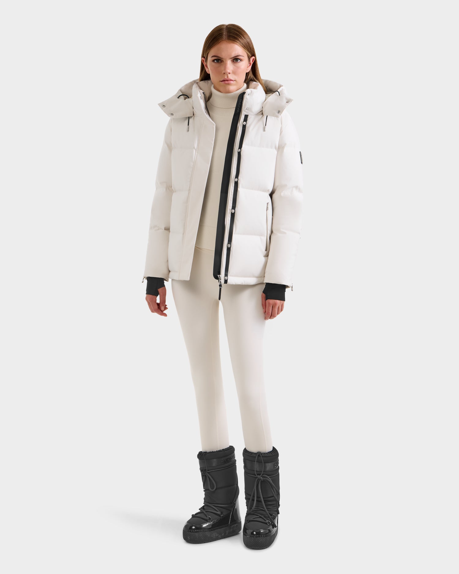 A woman faces forward, wearing the JOON H - 8125586 Vanilla quilted winter jacket with hood by Rudsak over a white turtleneck and leggings, paired with black winter boots, against a plain light background.