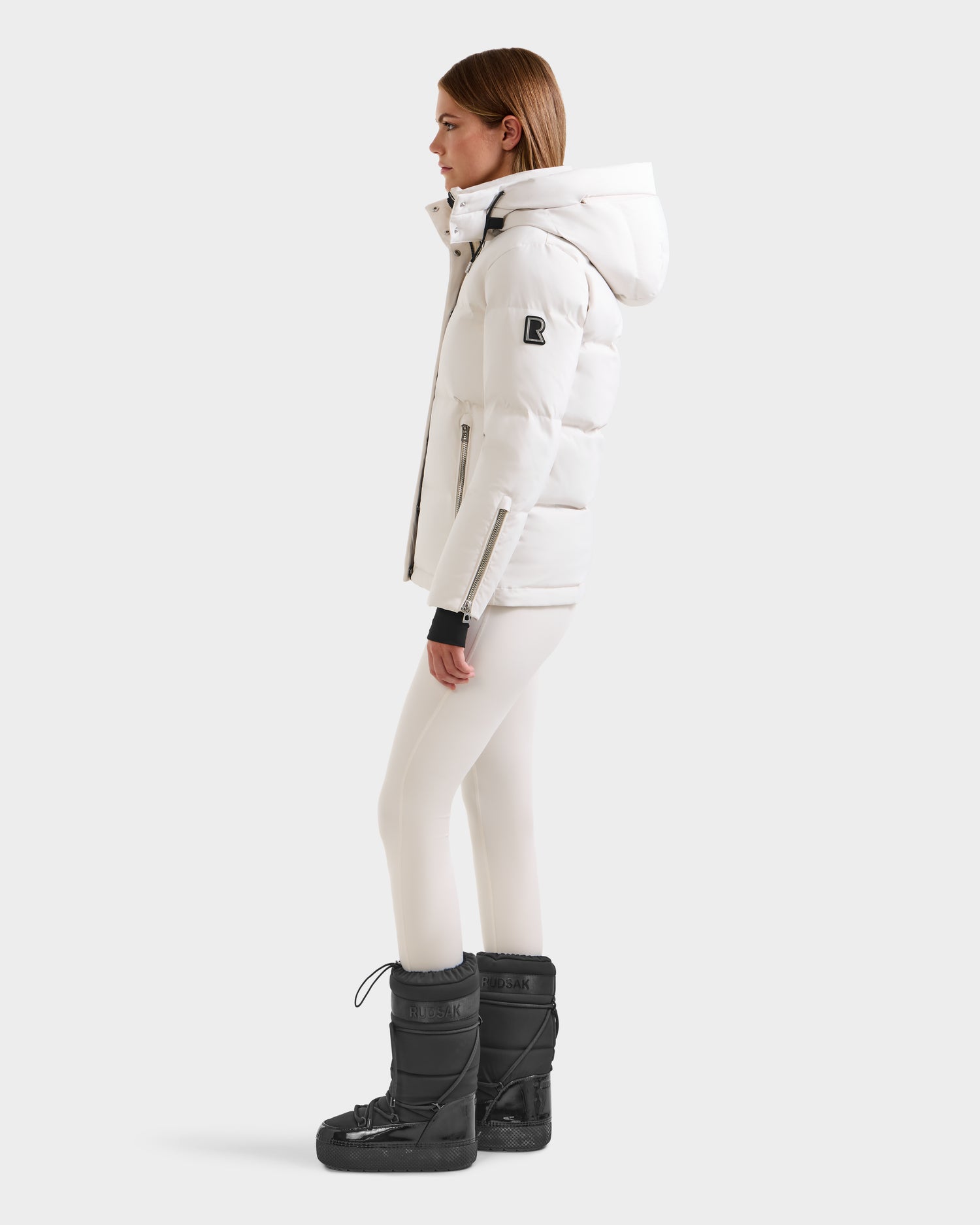 A woman stands in profile wearing the Rudsak JOON H - 8125586 Vanilla white quilted winter jacket with a hood, paired with white leggings and black winter boots against a plain light background.