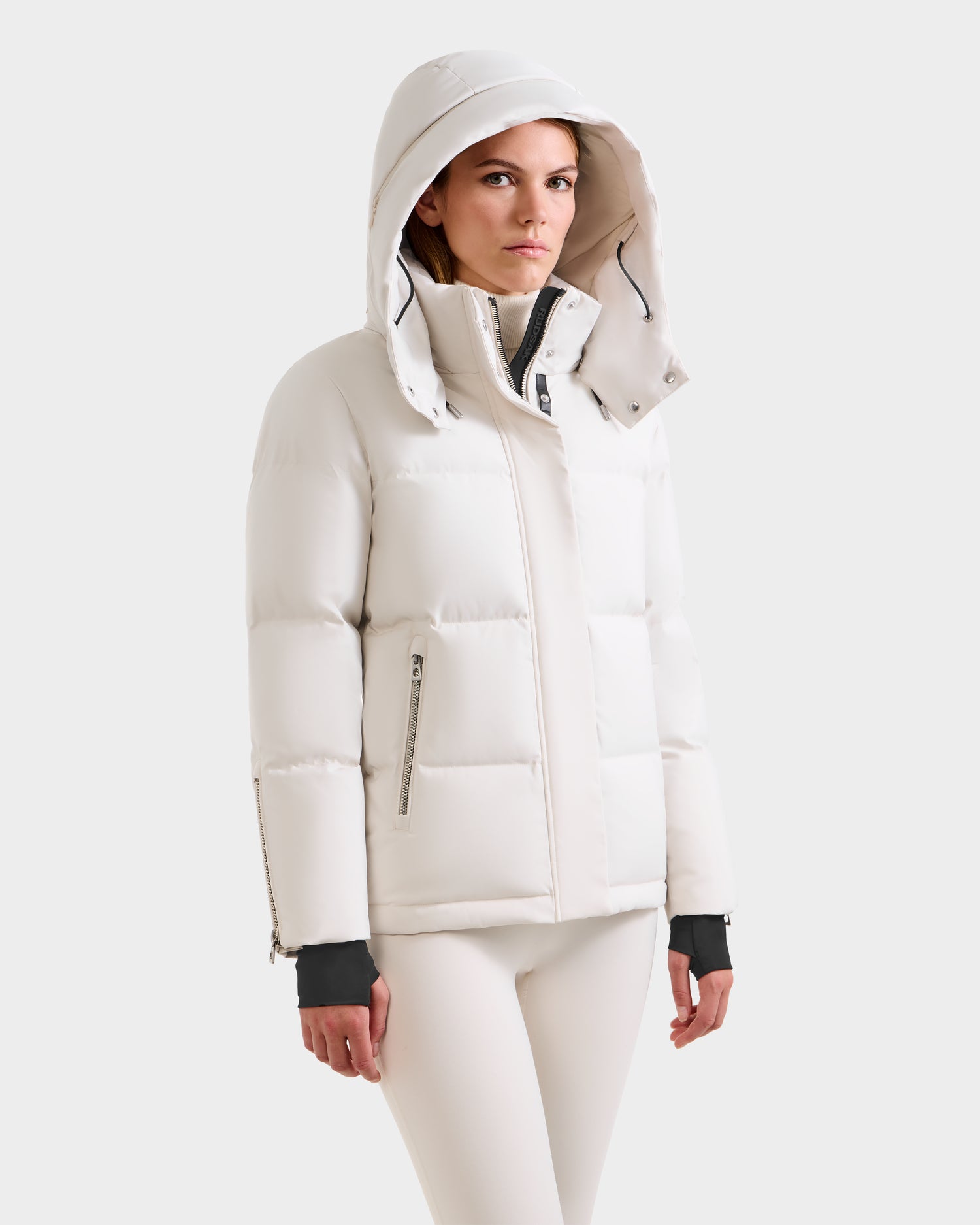 A woman models the Rudsak JOON H - 8125586 Vanilla quilted winter jacket with a hood, paired with light-colored pants, standing against a plain light gray background and looking straight ahead with a neutral expression.