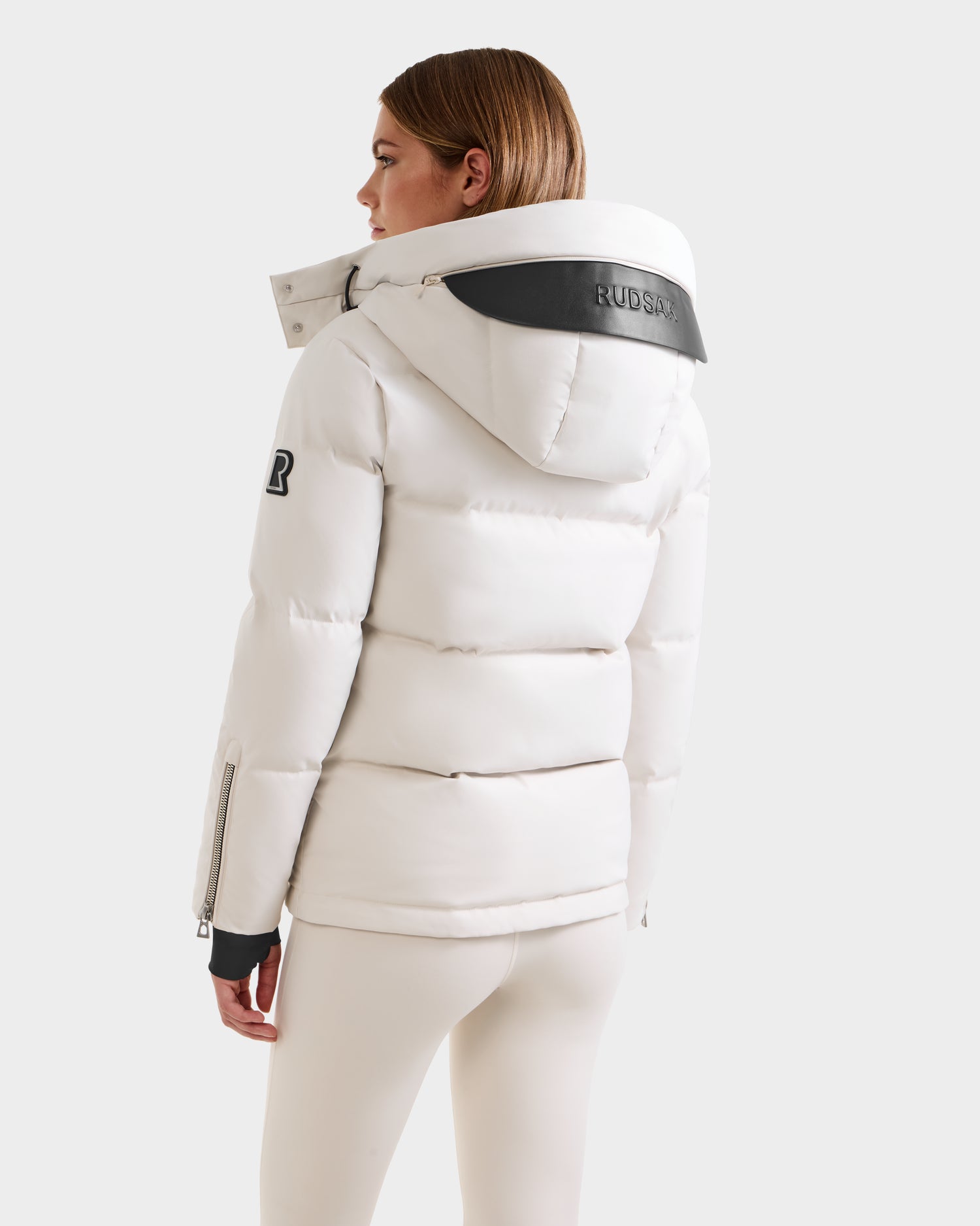 A woman stands facing away, wearing the Rudsak JOON H - 8125586 Vanilla quilted winter jacket with a large hood, black collar detailing, sleeve patch, and cuff zipper, paired with white leggings. The down jacket is made from recycled shell.