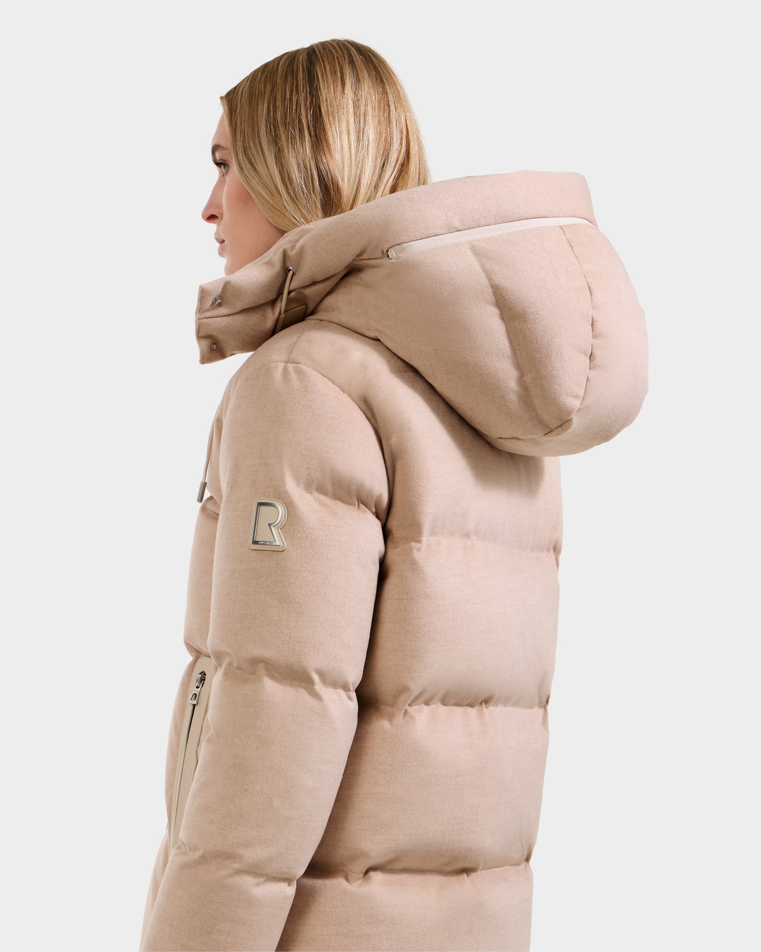 A blonde woman wears the Rudsak JOON W - 8125566 Heather Beige wool-blend down puffer, featuring a large hood, logo patch on the sleeve, and zippered pockets, shown from the side and back on a white background.