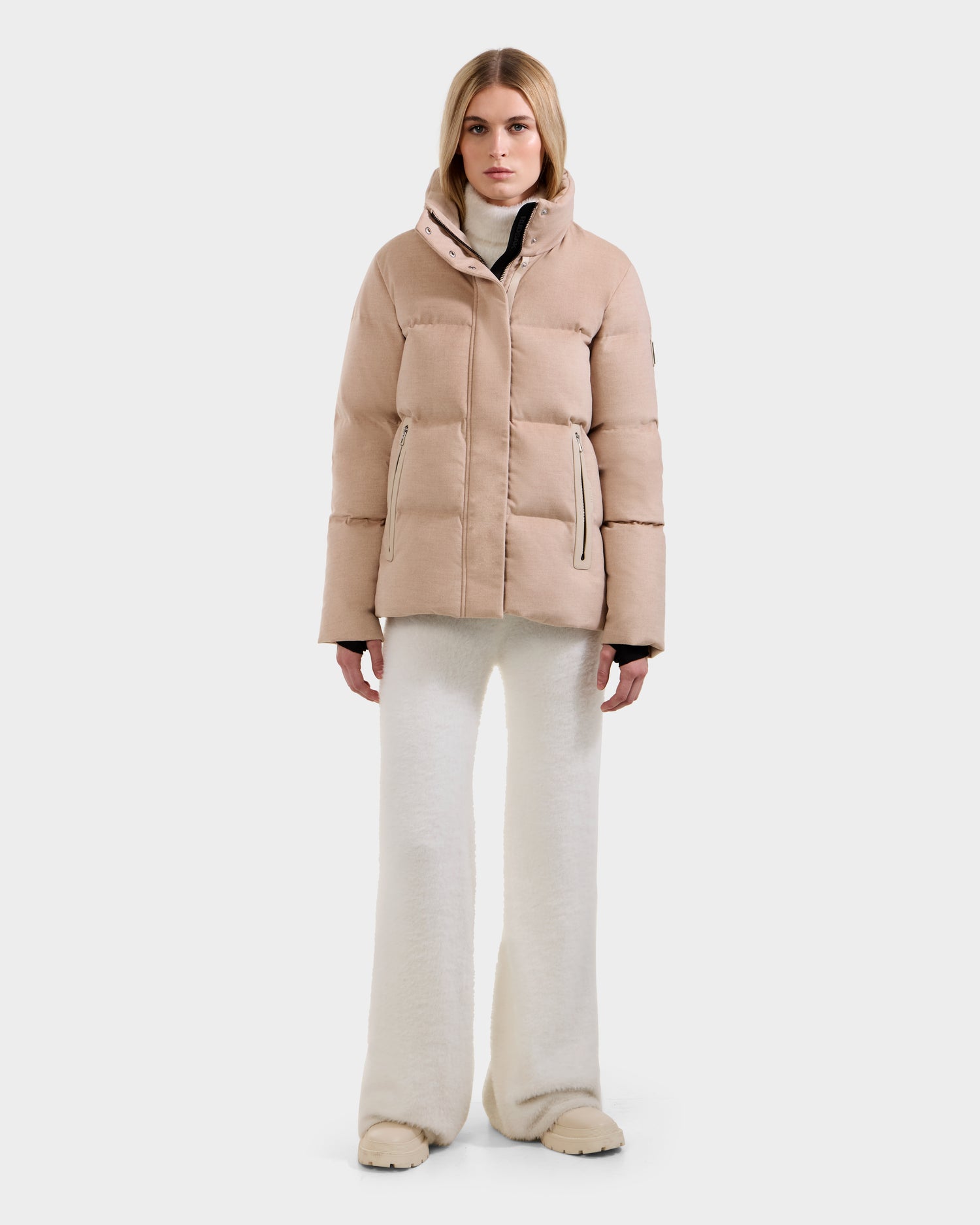 A woman stands facing forward against a plain white background, wearing the JOON W - 8125566 Heather Beige lightweight down jacket by Rudsak, with white wide-leg pants and beige shoes.