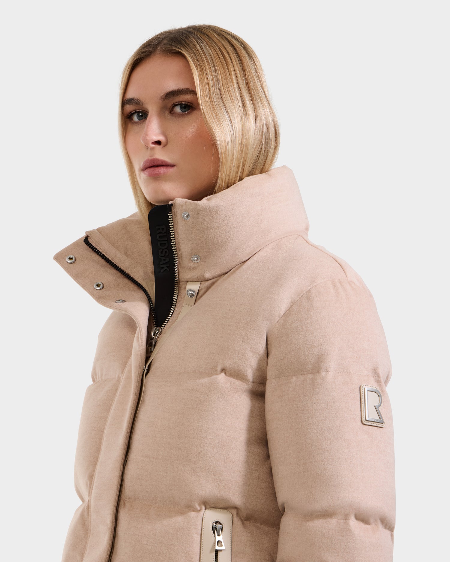 A woman with straight blonde hair wears the Rudsak JOON W - 8125566 lightweight down jacket in heather beige. Featuring a high collar, zip closures, and a sleeve logo patch, she looks calmly at the camera against a plain light background.