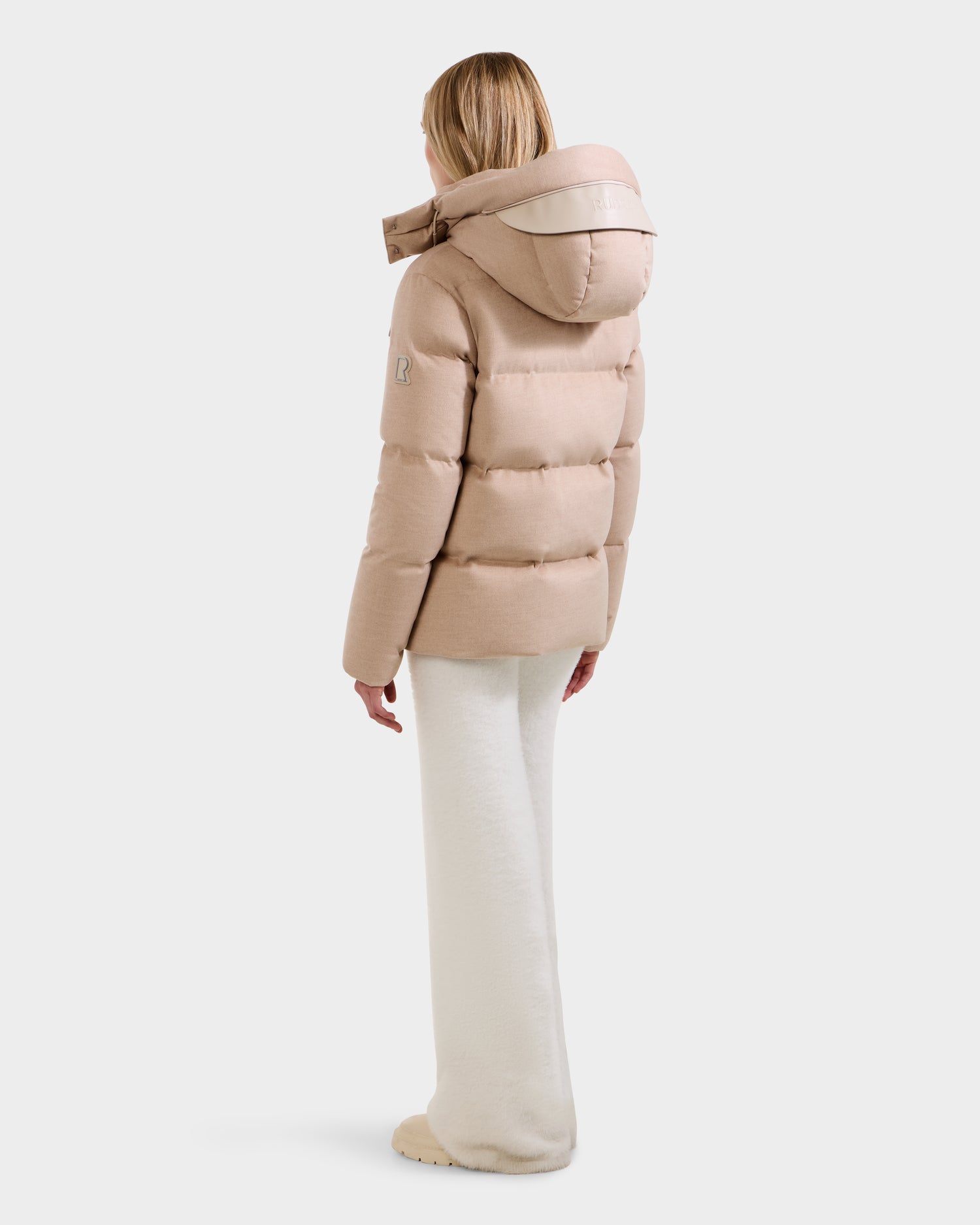 A person faces away, wearing the Rudsak JOON W - 8125566 Heather Beige women’s wool-blend puffer with a large hood and cream wide-leg pants. The light beige down jacket stands out against the plain white background.