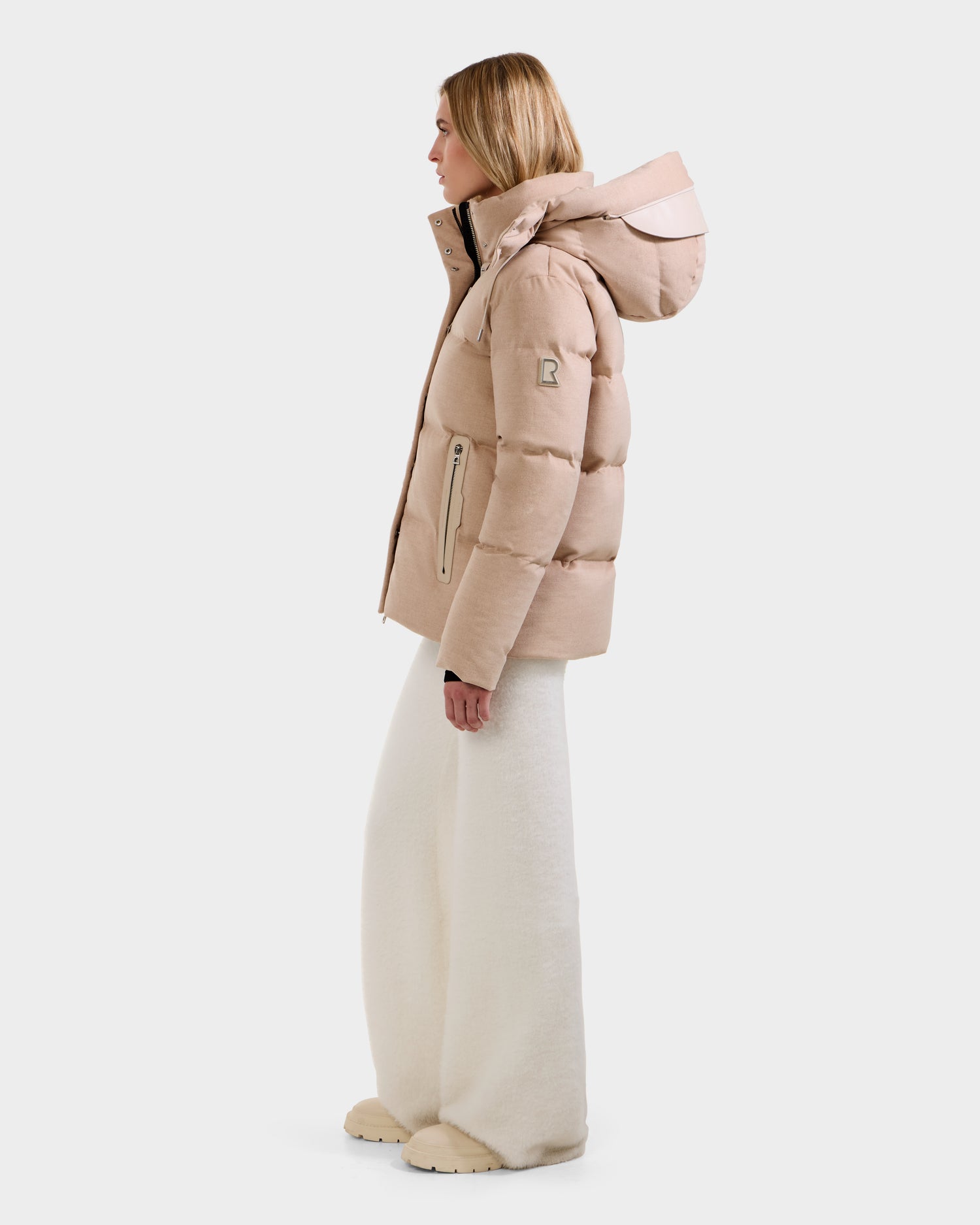 A woman stands in profile against a white background wearing the Rudsak JOON W - 8125566 Heather Beige women’s wool-blend down puffer with a hood, paired with wide-leg white pants and beige shoes.