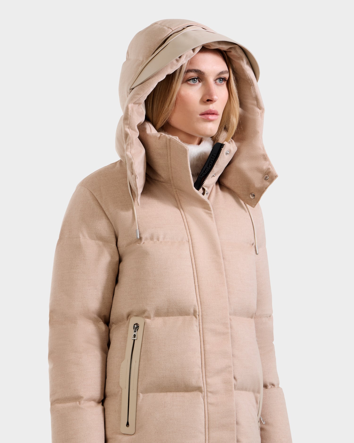 A woman wearing the Rudsak JOON W - 8125566 Heather Beige wool-blend down puffer with a hood, zippered pockets, and high collar stands against a plain white background, looking slightly to the side.