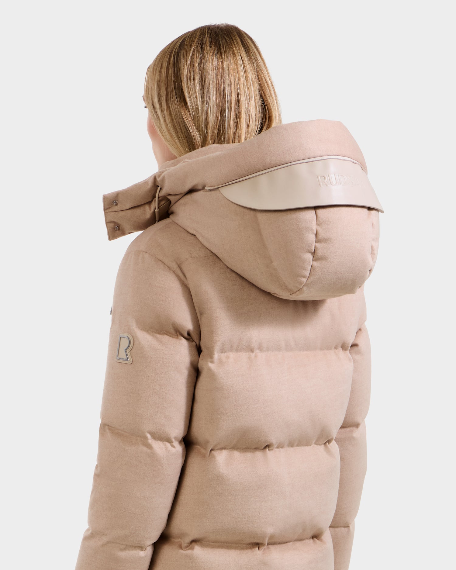A person with light brown hair is seen from the back wearing the Rudsak JOON W - 8125566 Heather Beige women’s wool-blend down puffer with a hood, logo patch on the sleeve, and “RUDSAK” text accent on the hood.