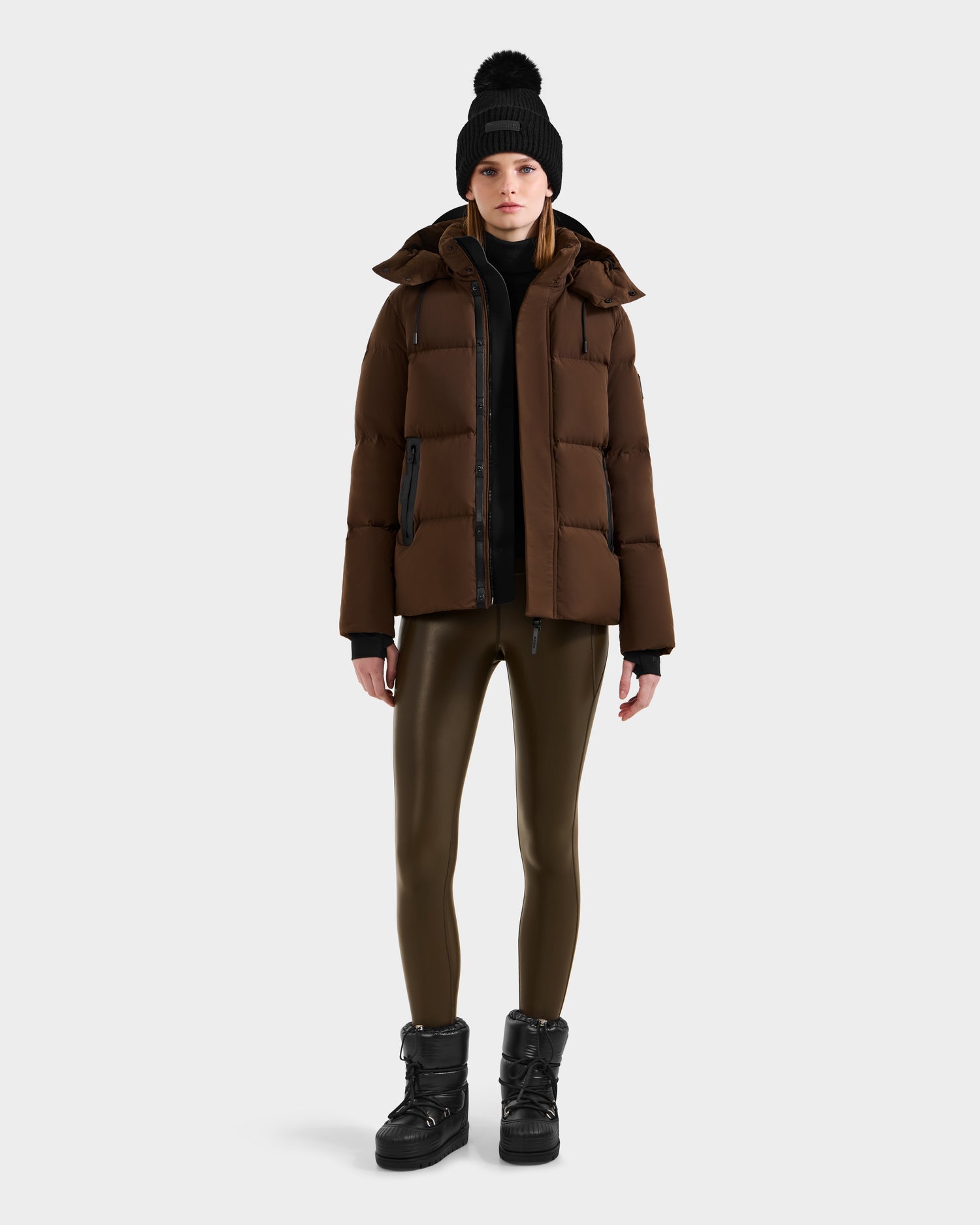 A person wearing the Rudsak JOON - 8124728 Mocha puffer jacket stands facing forward against a plain white background, styled with a black beanie, black top, shiny olive-green leggings, and black winter boots.