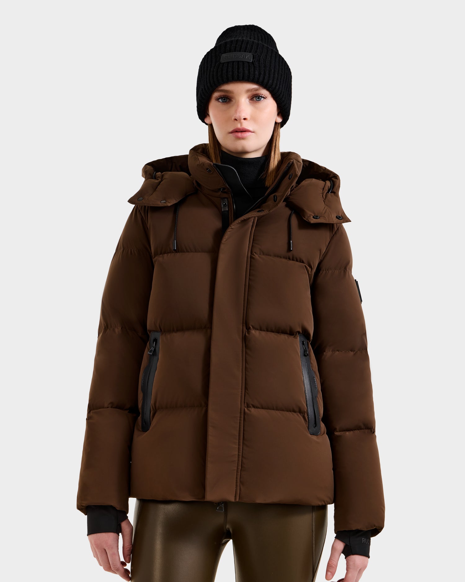 A person wearing the Rudsak JOON - 8124728 Mocha brown down puffer jacket with a hood, a black beanie, black turtleneck, and shiny brown pants stands facing forward against a plain light gray background.