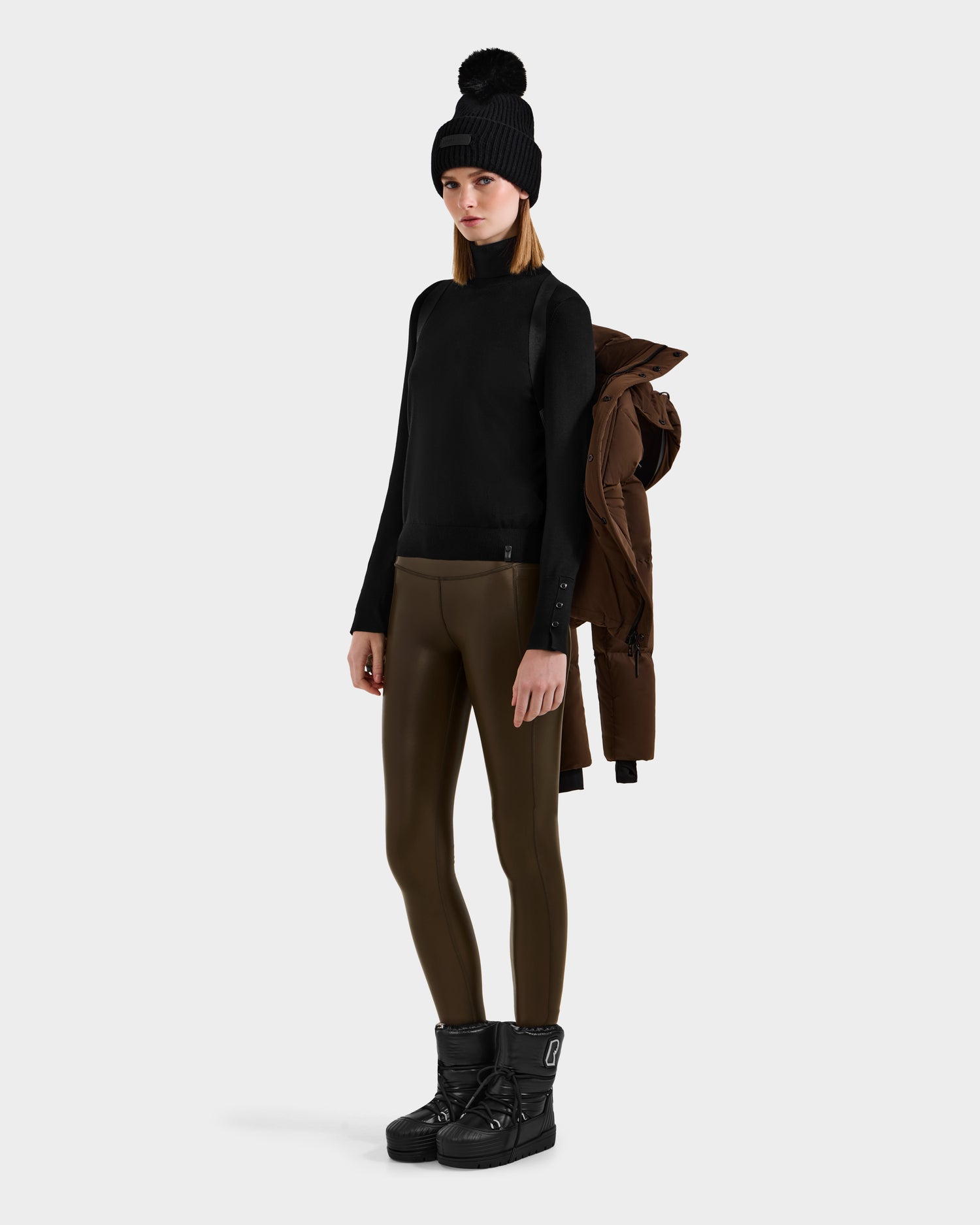 A woman in a black beanie, black sweater, olive green leggings, and black winter boots holds the Rudsak JOON - 8124728 Mocha lightweight down puffer jacket over her shoulder against a plain light background.