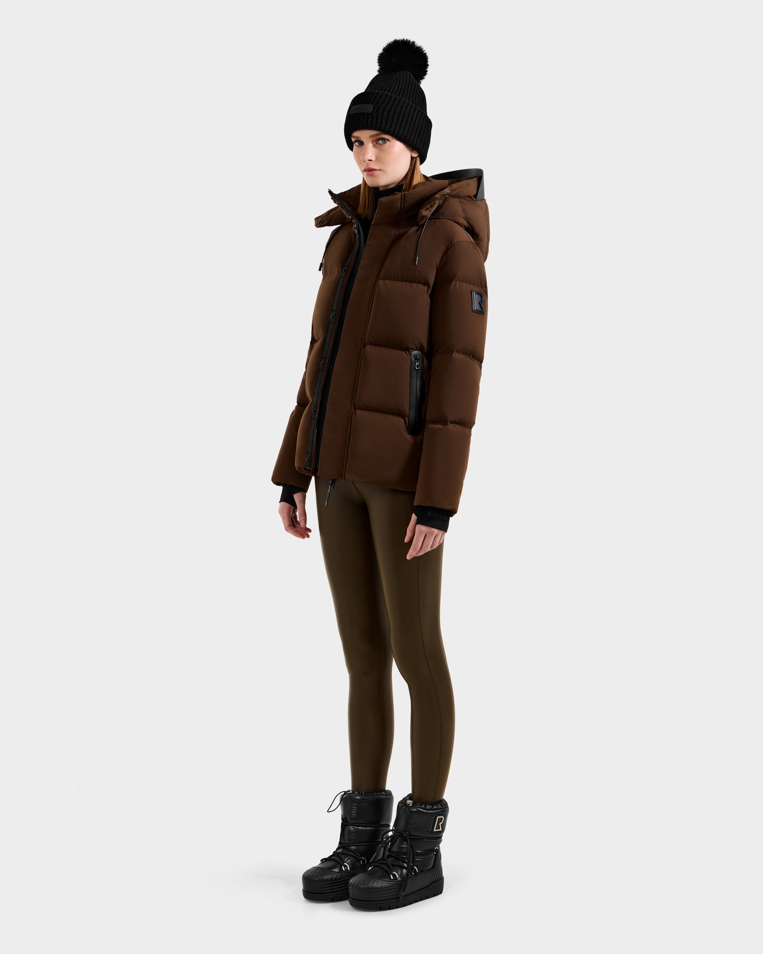 A person wearing the Rudsak JOON - 8124728 Mocha brown down puffer jacket, a black pom-pom beanie, olive leggings, and black winter boots stands against a plain light gray background.