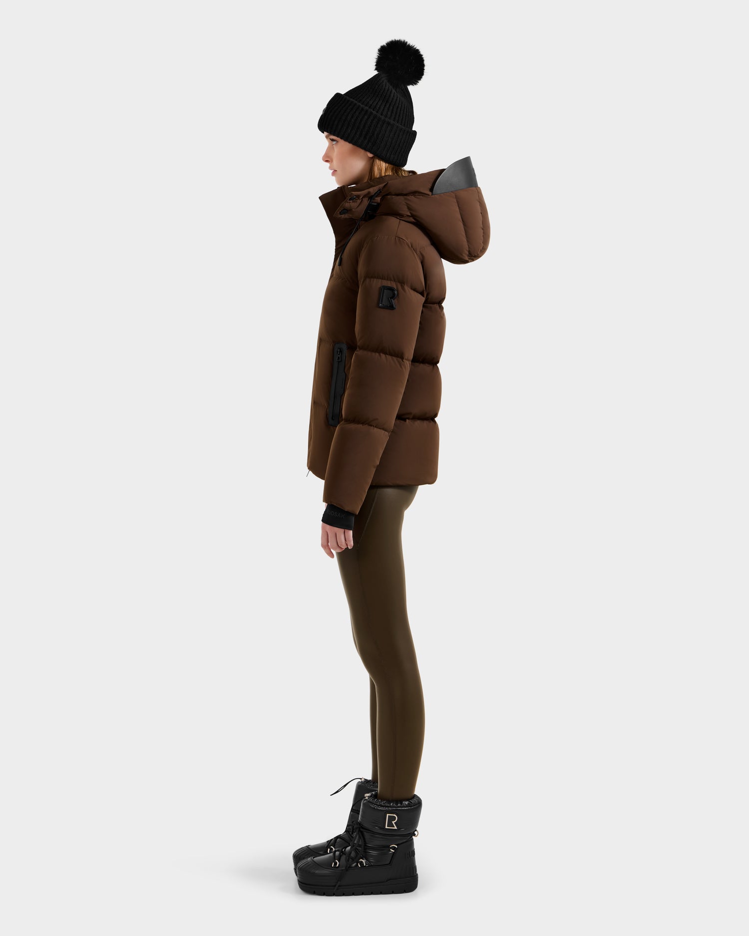 A woman in side profile wears the Rudsak JOON - 8124728 Mocha down puffer jacket with a hood, paired with olive leggings, black winter boots, and a black pom-pom beanie, standing against a plain light gray background.