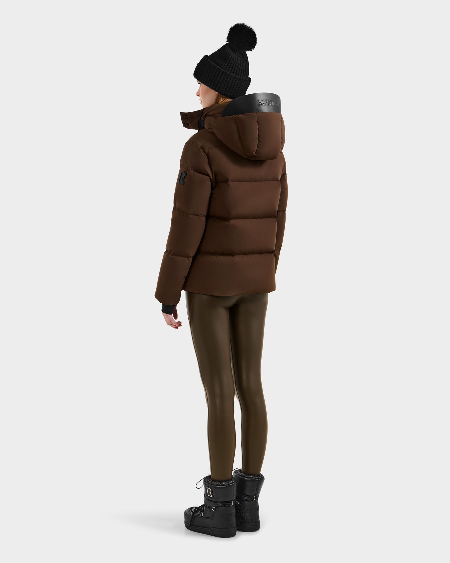 A person wearing the Rudsak JOON - 8124728 Mocha puffer jacket, a black pom-pom beanie, shiny olive leggings, and black winter boots stands facing away on a plain light background.