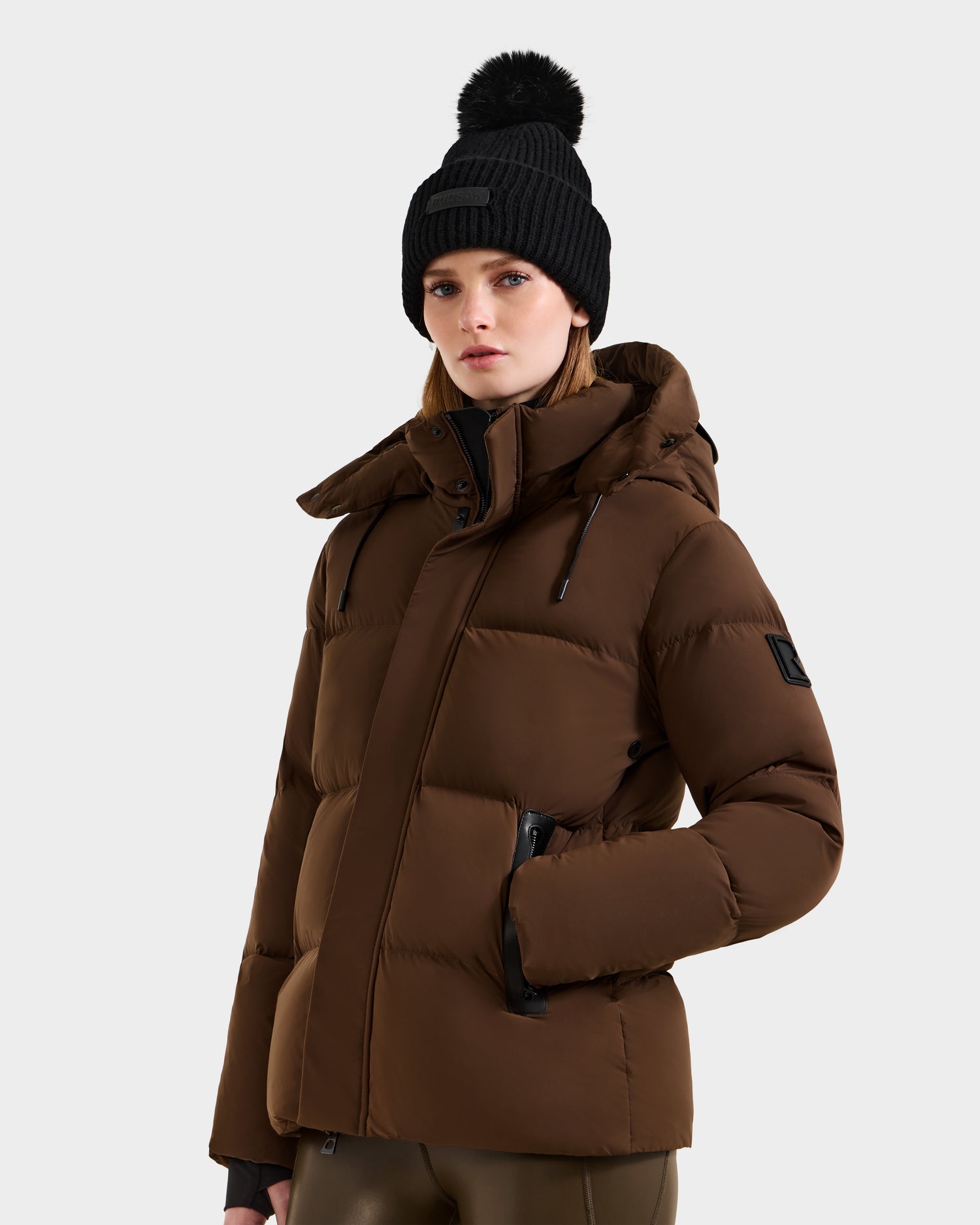 A woman stands facing forward in a Rudsak JOON - 8124728 Mocha down puffer jacket with hood and black zipper details, paired with a black knit beanie featuring a pom-pom. The background is plain and light-colored.