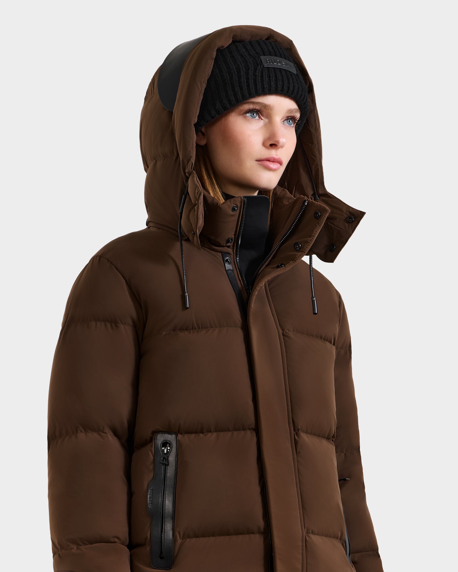 A person in the Rudsak JOON - 8124728 Mocha puffer jacket with a hood and a black knit beanie, looking slightly to the side against a plain light background.