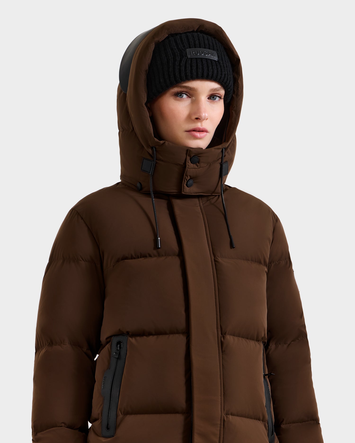 A woman in the Rudsak JOON - 8124728 Mocha down puffer jacket with a hood and a black knit beanie stands against a plain light background.