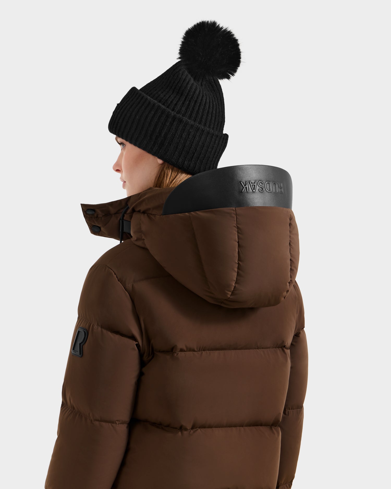 A person in the Rudsak JOON - 8124728 Mocha brown down puffer jacket with a black sleeve patch and hood, paired with a black pom-pom beanie, is seen from the side and back against a plain light background.