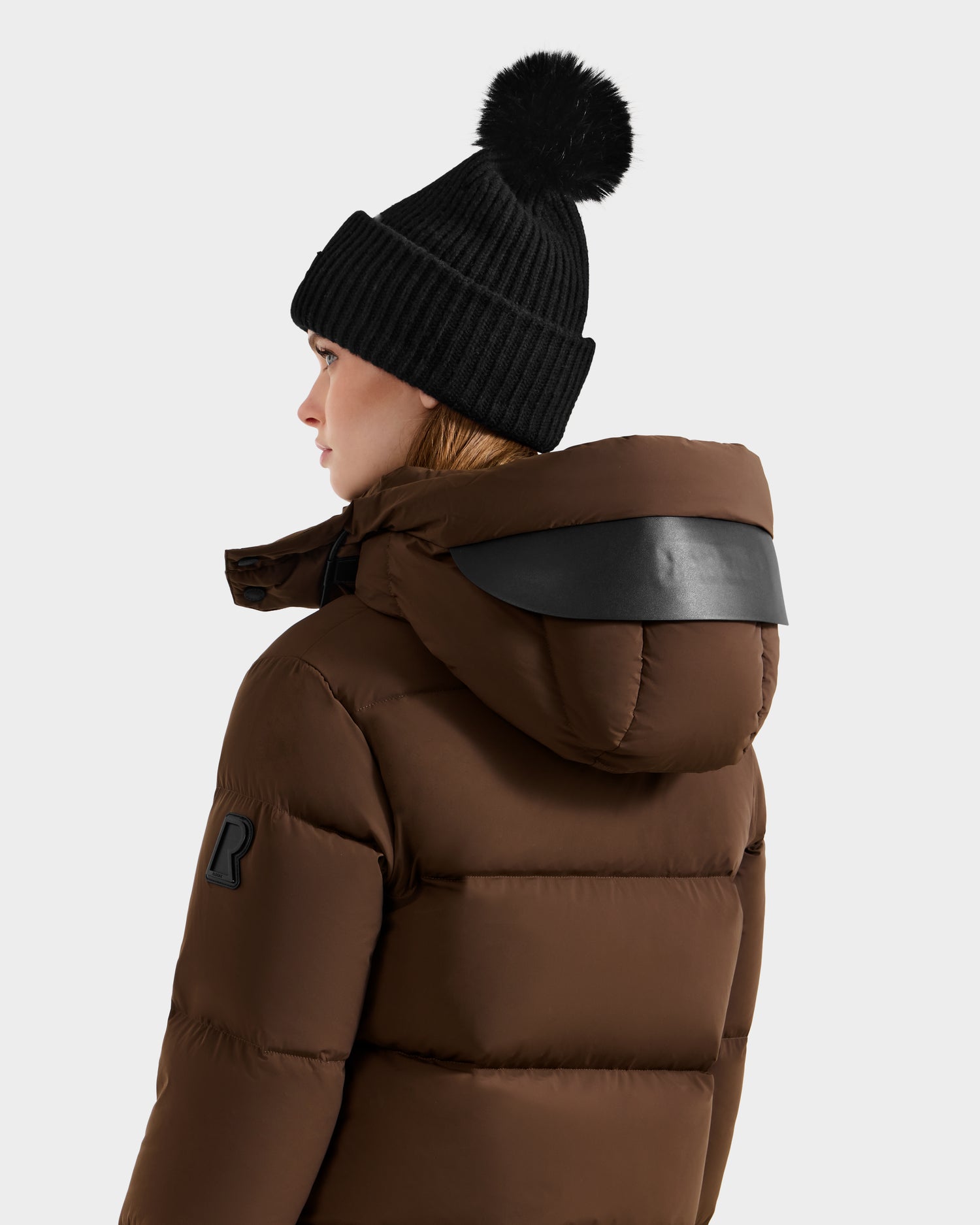 Someone wearing the Rudsak JOON - 8124728 Mocha brown down puffer jacket and a black knit pom-pom beanie, seen from behind against a plain light background.
