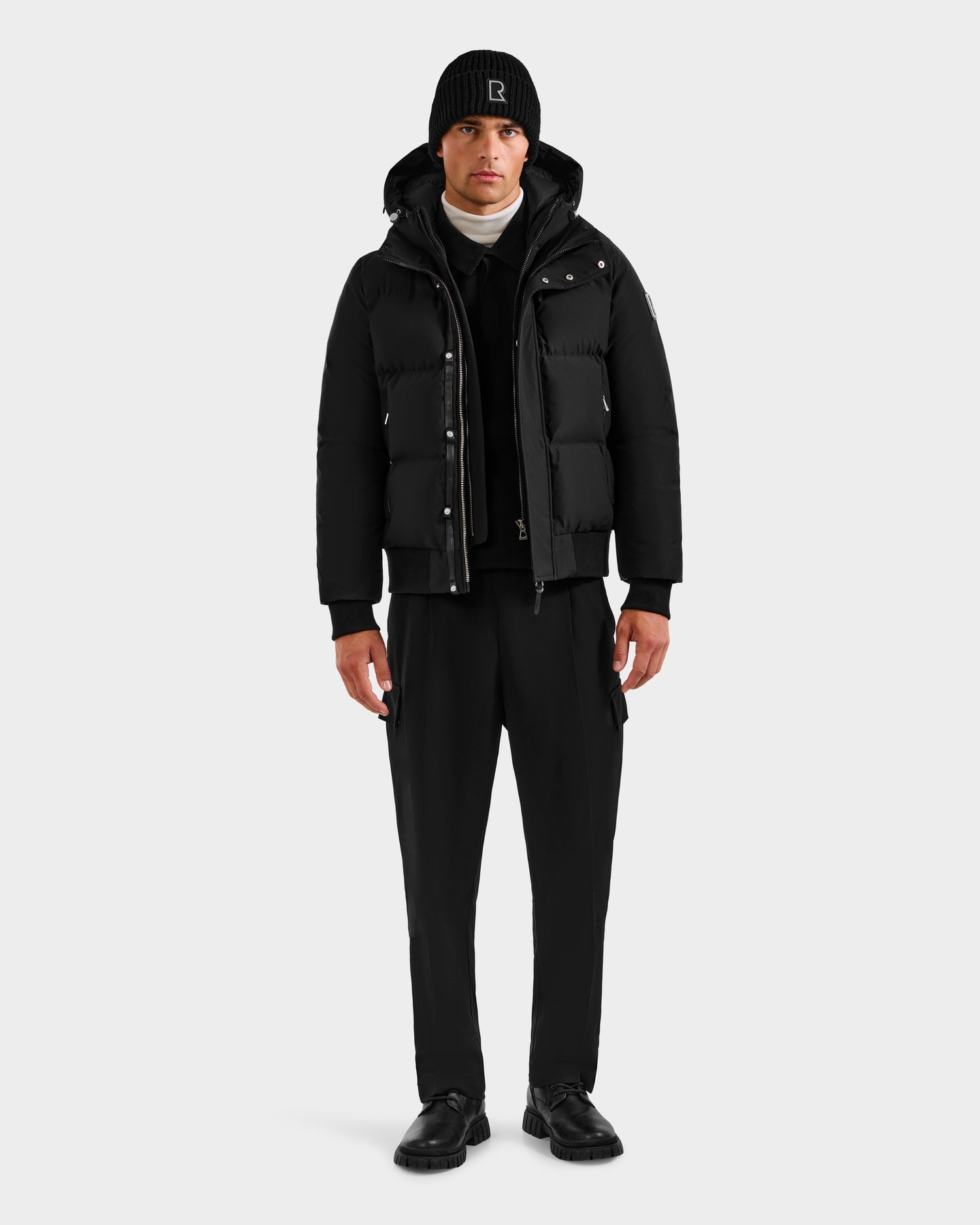 A man faces forward against a plain light gray background, wearing the Rudsak JORDY H - 6125689 Black quilted down bomber jacket, paired with black pants, black beanie, black shoes, and a white turtleneck sweater for winter warmth.