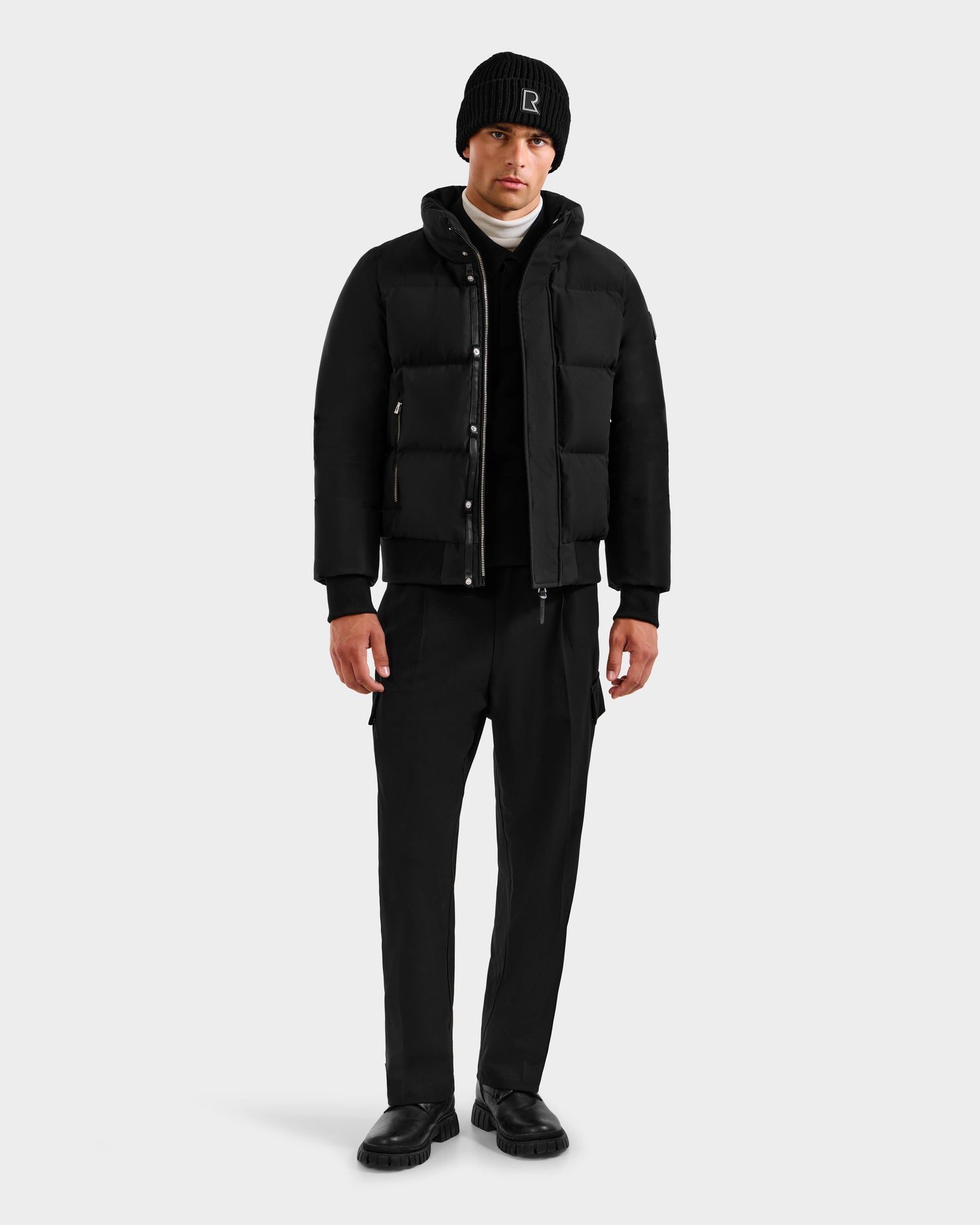 A man faces forward against a light gray background, wearing the Rudsak JORDY H - 6125689 Black quilted down bomber puffer jacket, black pants and shoes, a white turtleneck, and a logo beanie, showcasing stylish winter warmth.