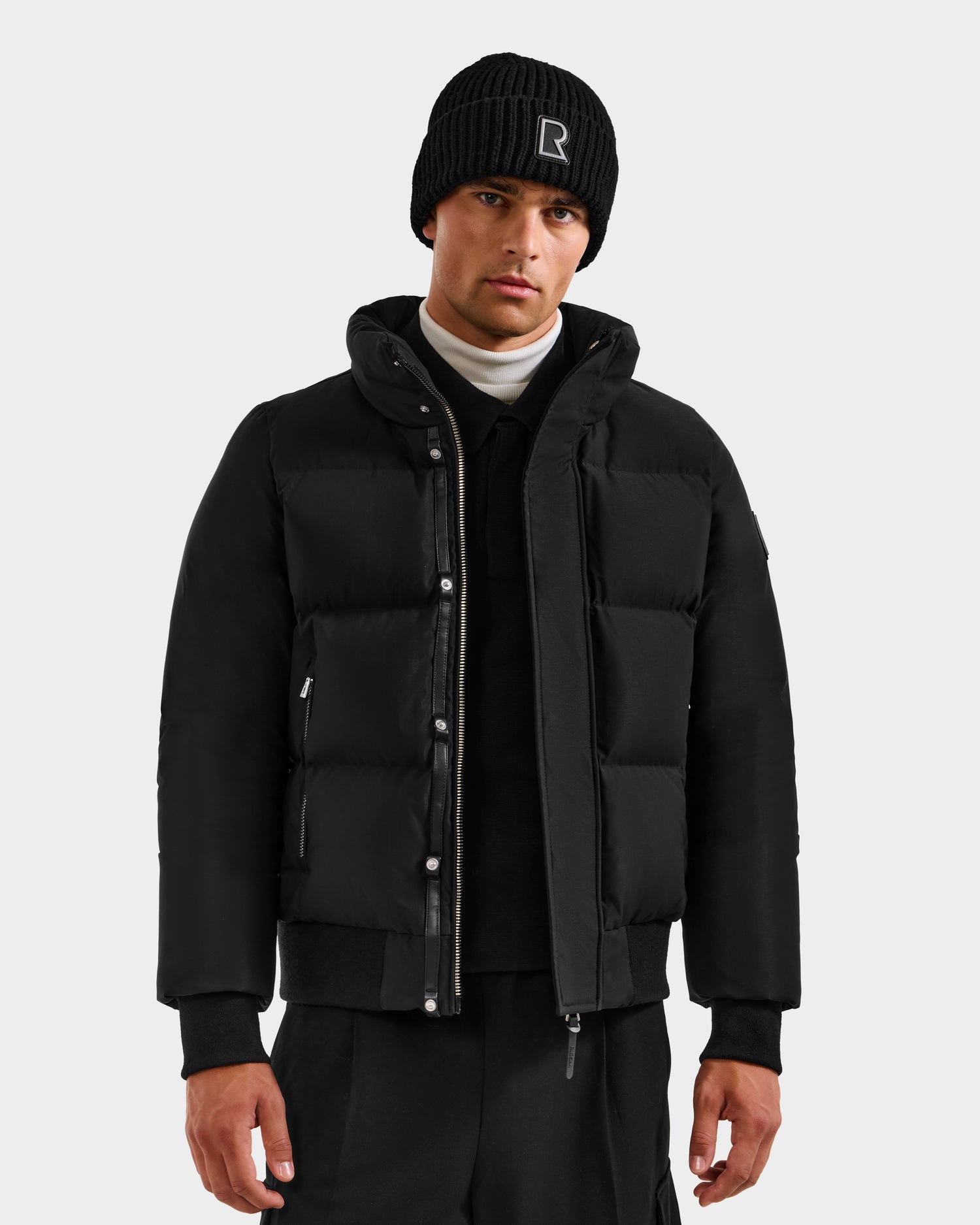A man stands facing forward against a plain light gray background, wearing the Rudsak JORDY H - 6125689 Black puffer jacket, a black knit beanie with an R, black pants, and a white turtleneck.