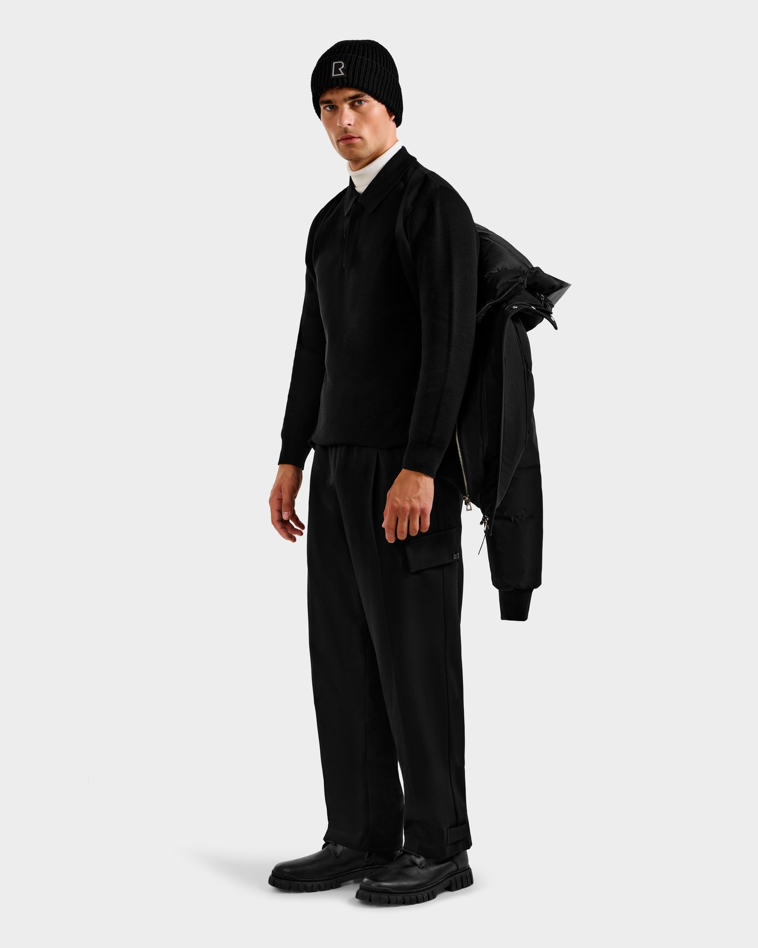 A man stands against a plain light background, dressed in all black with chunky shoes, a knit beanie, and the Rudsak JORDY H - 6125689 Black men’s quilted down bomber jacket draped over his shoulder.