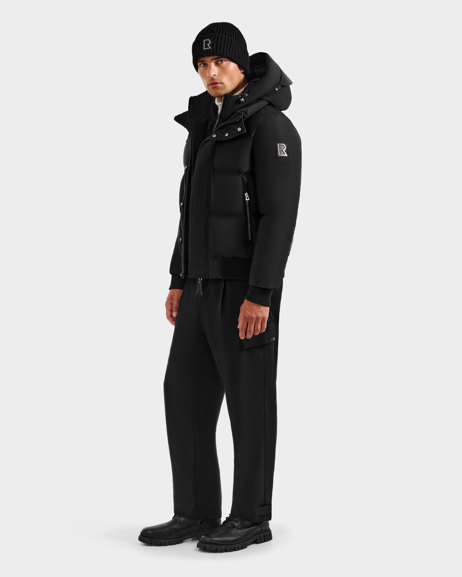 A person wears the Rudsak JORDY H - 6125689 Black men’s quilted down bomber with black sweatpants, a knit beanie, and shoes against a white background—ideal winter style.