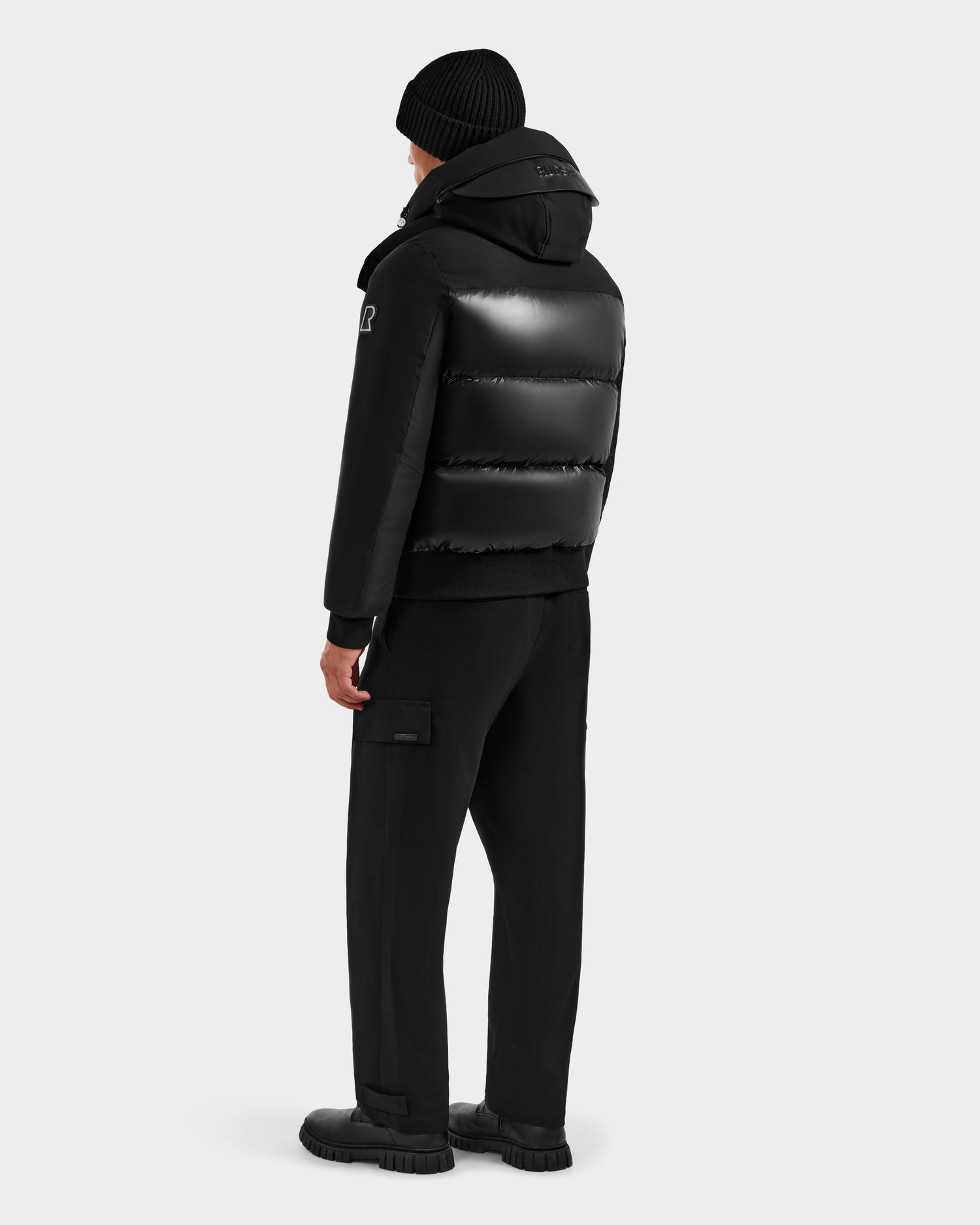 A person faces away wearing the Rudsak JORDY H - 6125689 Black quilted down bomber, paired with black cargo pants and shoes, ready for winter warmth against a plain light gray background.