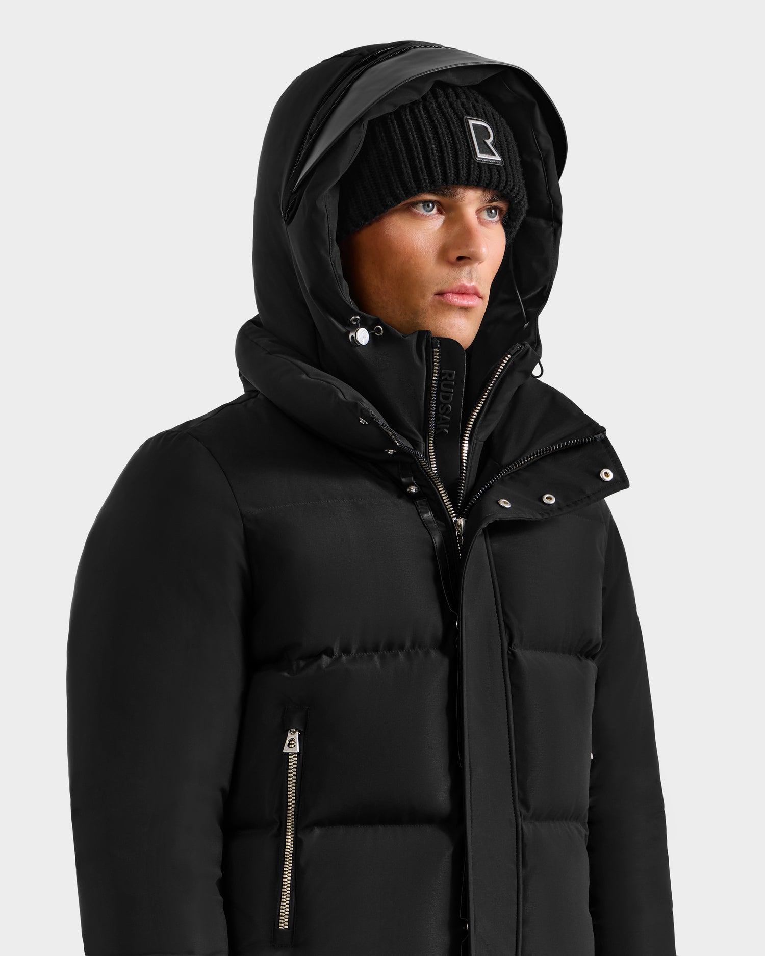 Wearing the Rudsak JORDY H - 6125689 Black men's quilted down bomber with hood and a black beanie, this person exudes effortless winter warmth against a plain light background.