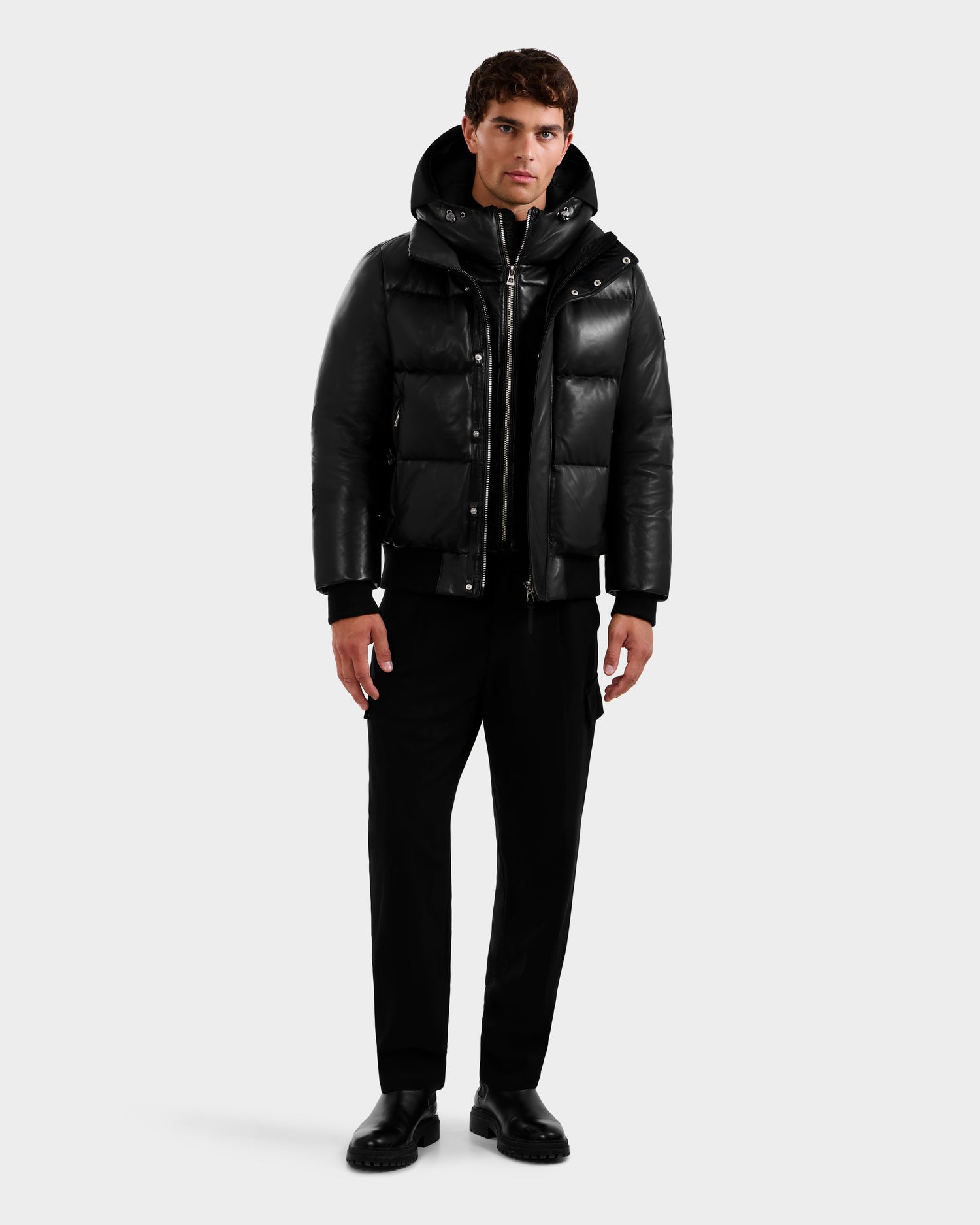 A man faces forward wearing the Rudsak JORDY X - 6125694 Black quilted bomber over a hoodie, with black pants and black shoes, set against a plain, light-colored background.