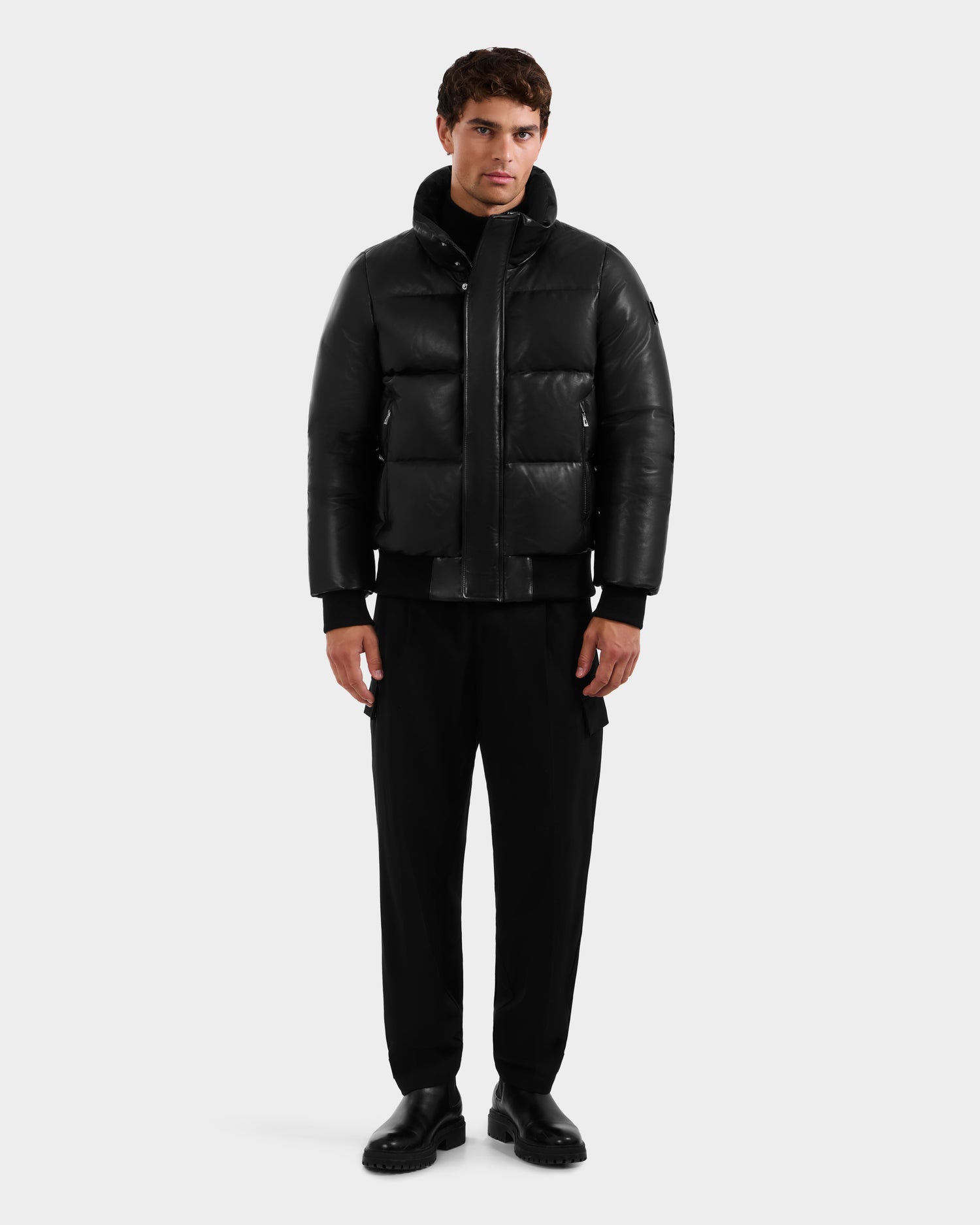 A man faces forward with a neutral expression, wearing the Rudsak JORDY X - 6125694 Black quilted bomber jacket, black pants, and black shoes, set against a plain light gray background.