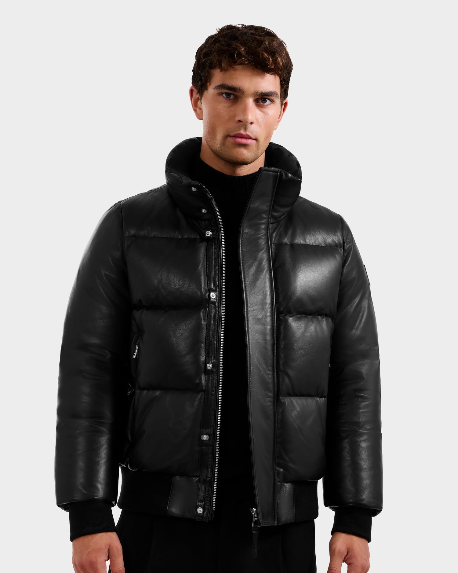 A man with short curly brown hair and a neutral expression faces the camera, wearing the Rudsak JORDY X - 6125694 Black quilted bomber over a black shirt, partially unzipped, against a plain light gray background.