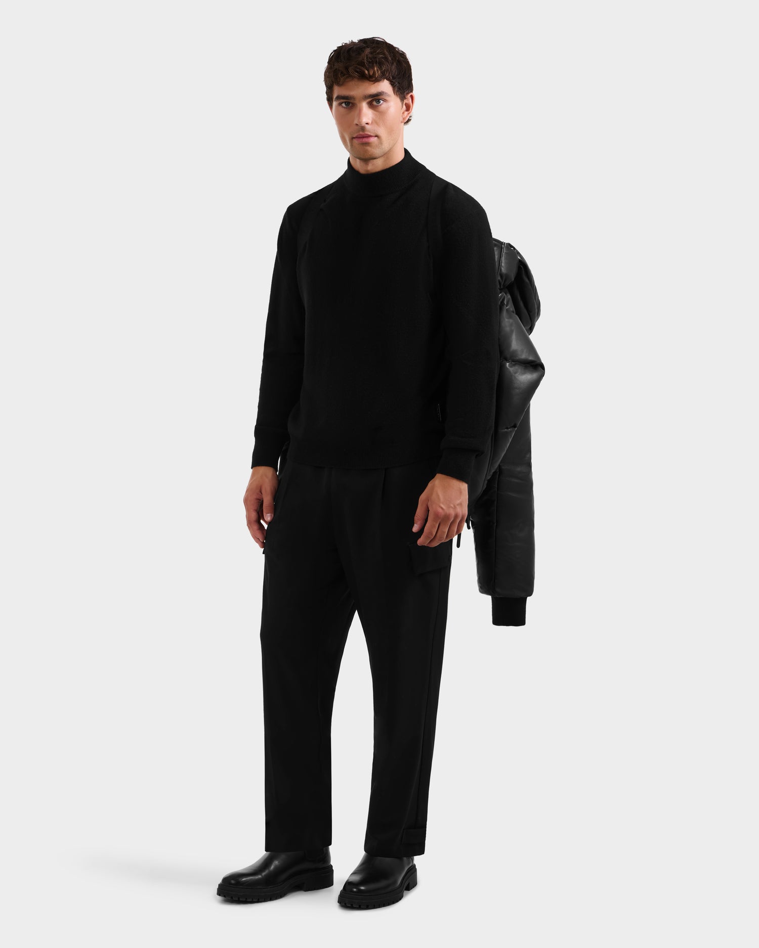A man stands against a plain light gray background, wearing the Rudsak JORDY X - 6125694 Black unisex bomber draped over his left shoulder, paired with black sweater, black pants, and black shoes.