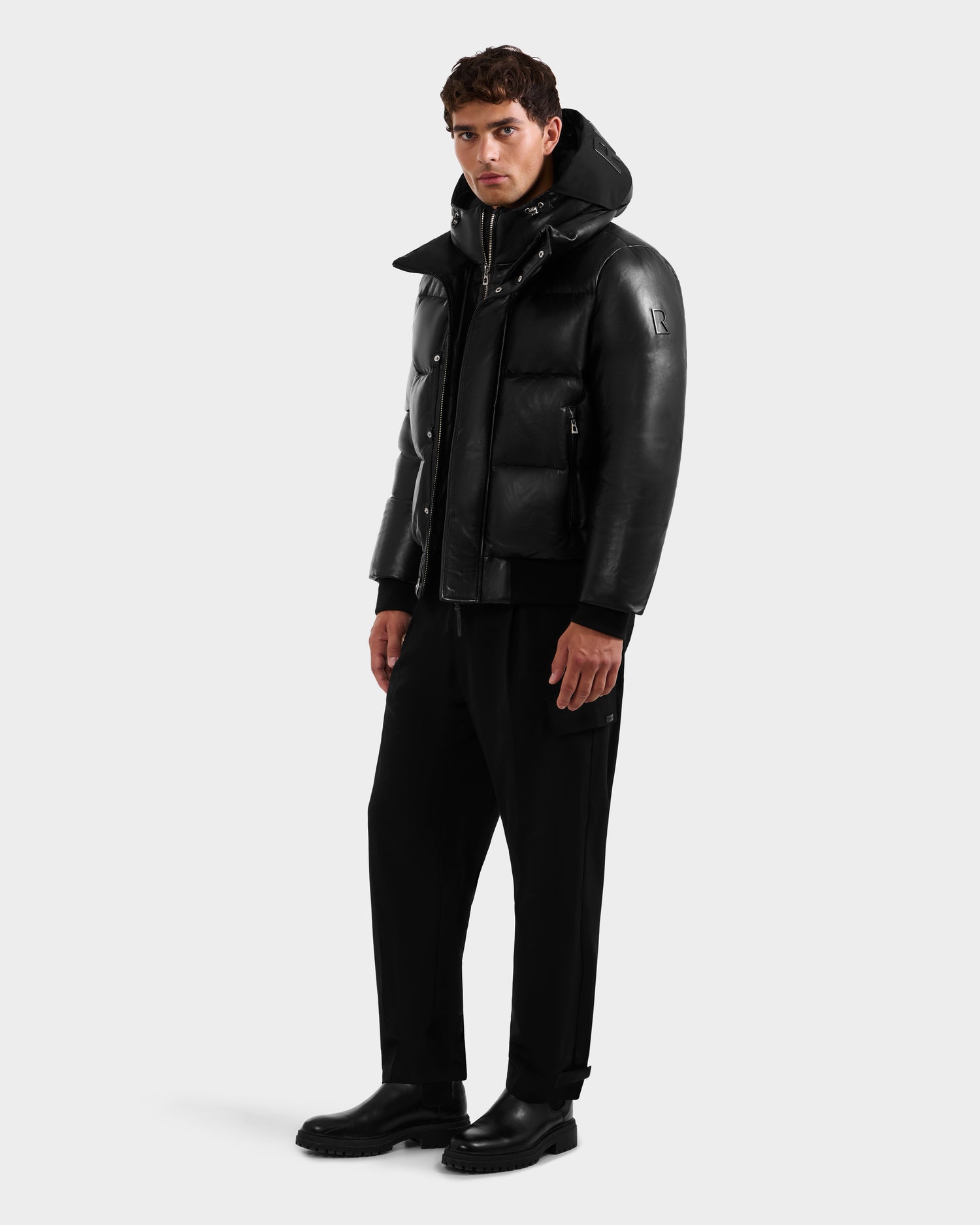 A man stands facing forward against a plain background, wearing the Rudsak JORDY X - 6125694 Black quilted bomber jacket with a hood, paired with black pants and boots. His expression is neutral and his arms are at his sides.