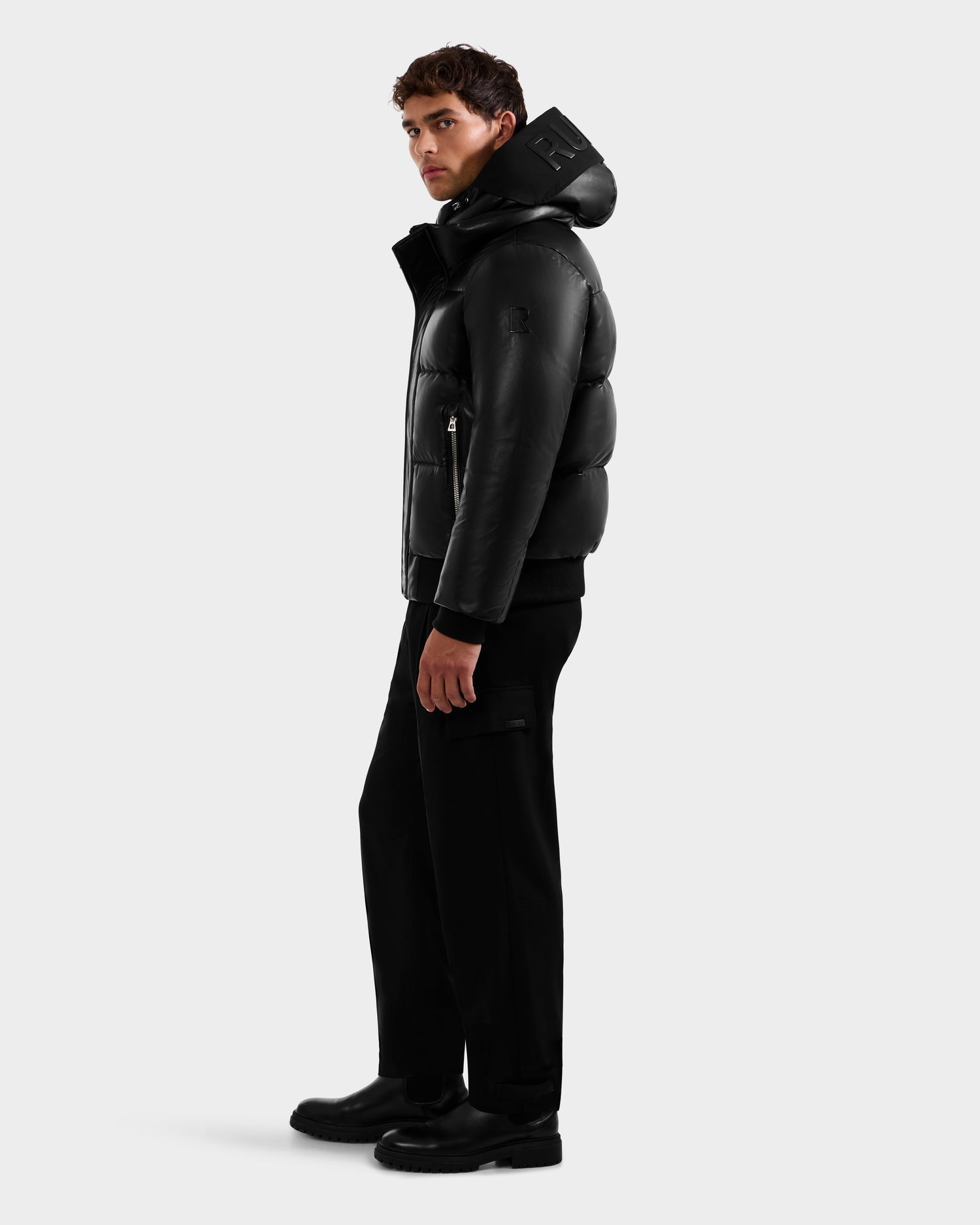 A man stands sideways against a white background, wearing the Rudsak JORDY X - 6125694 Black quilted bomber jacket with a hood, paired with black pants and black shoes.