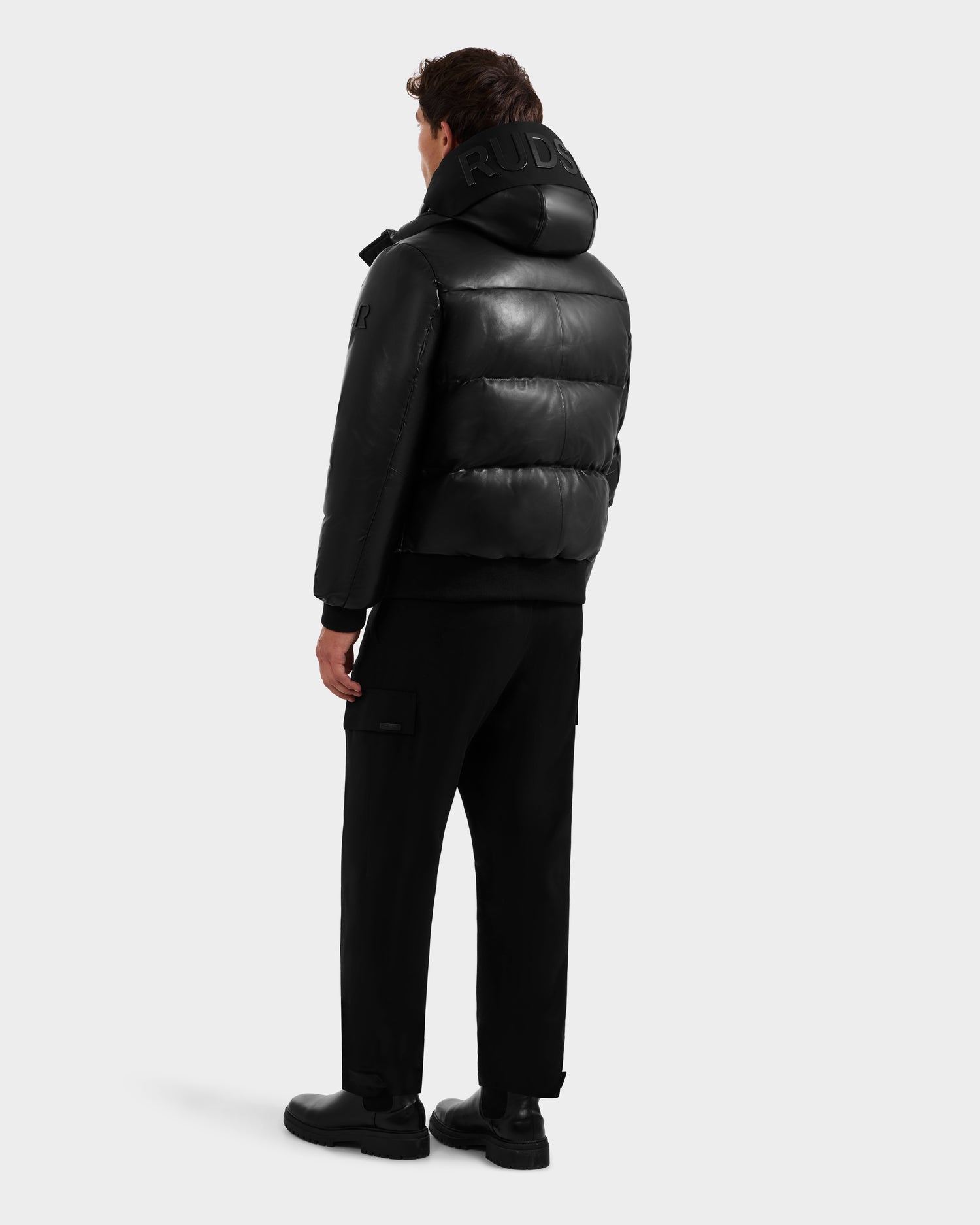A man faces away wearing the Rudsak JORDY X - 6125694 Black quilted bomber with a large hood, paired with black pants and shoes, set against a plain light gray background.