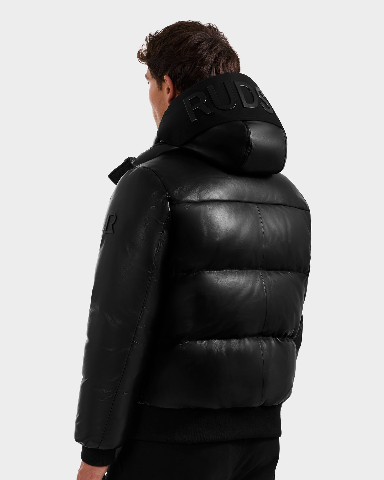 A man faces away wearing the Rudsak JORDY X - 6125694 Black bomber jacket with a large embossed hood. The shiny leather and modern design stand out against a plain light gray background.