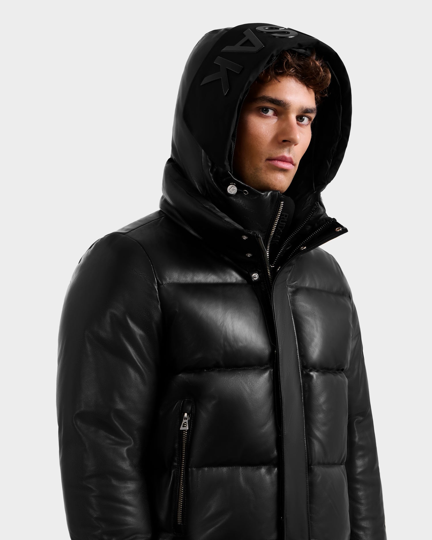 A young man with curly hair wears the Rudsak JORDY X - 6125694 Black quilted bomber puffer jacket with a hood, high collar, zippered pockets, and bold lettering along the hood edge, standing against a plain light background.
